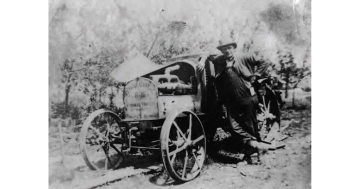 Black and white photo of a man leaning against a small wooden cart with large wheels, in an outdoor farm or rural area with trees and a fenced field in the background.