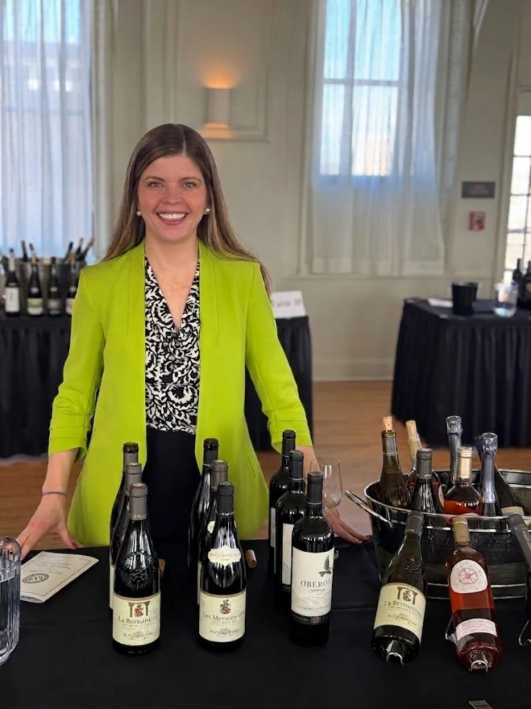 Woman smiling at a wine tasting event, standing behind a table with various wine bottles, bottles in buckets, and a glass of wine, in a well-lit room with windows and curtains.