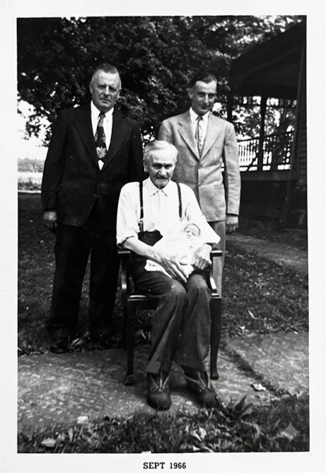 Three men, one seated with a baby on his lap, in a yard with trees and a porch in the background, September 1966.