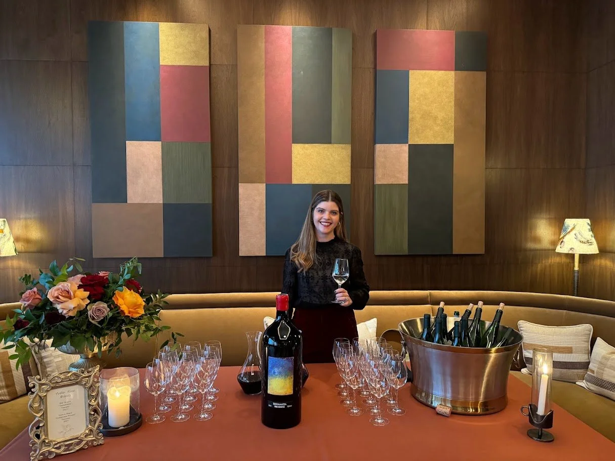 A woman in a black top standing behind a round table set for a wine tasting with a large bouquet of flowers, a candle, a bottle of wine, and wine glasses, in a cozy, warmly lit room with wooden walls and colorful abstract art.