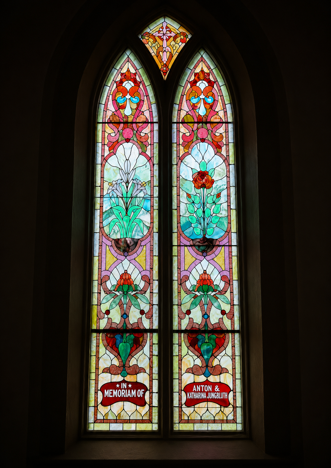 Colorful stained glass window with floral and abstract patterns, and a memorial plaque in the lower section that reads 'In Memoriam of Anton & Katharina Jungbluth'.