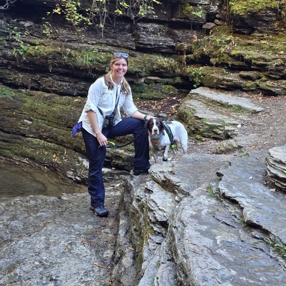 A woman smiling and kneeling on rocks with a dog in a natural outdoor setting with rock formations and greenery.