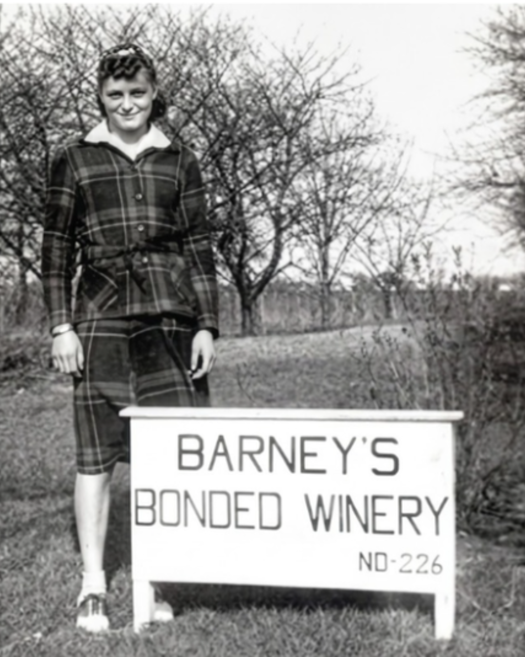 A young woman wearing a plaid dress with a belt, standing outdoors next to a sign that reads "Barney's Bonded Winery ND-226". Behind her, leafless trees are visible, indicating the photo was taken in a wooded or rural area.