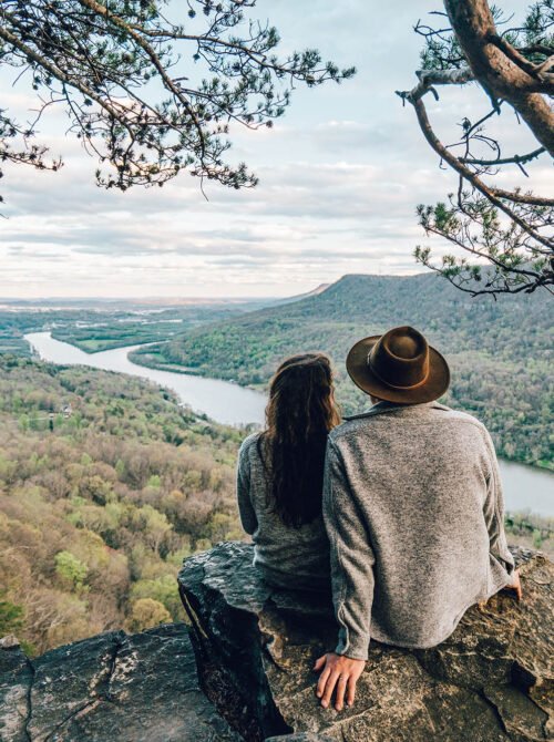 A man and woman sitting on a rocky ledge overlooking a river and forested landscape under a cloudy sky, with tree branches framing the scene.