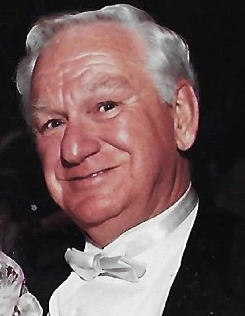 An elderly man with light skin, gray hair, dressed in formal attire including a black tuxedo and white bow tie, smiling at the camera.
