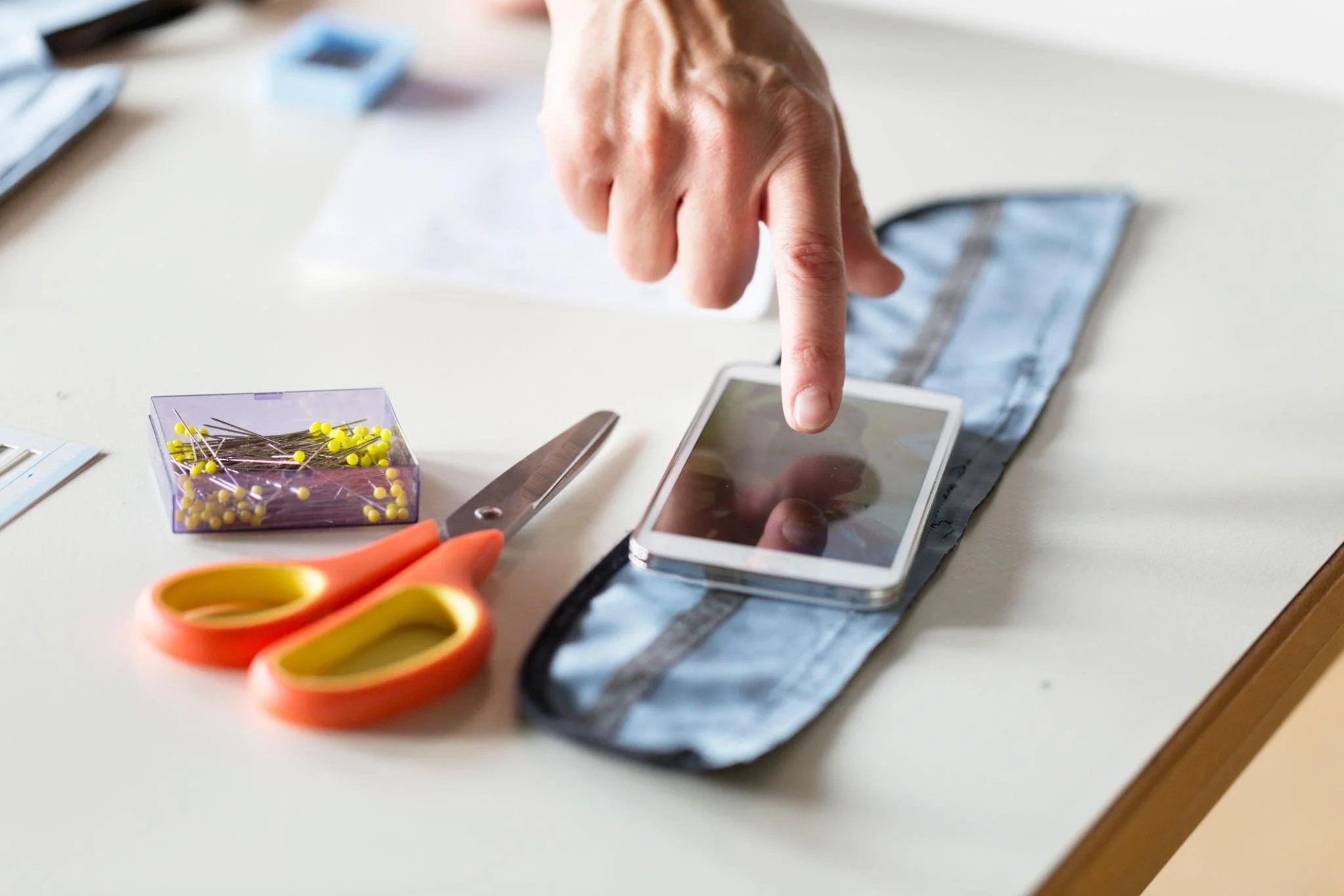 A person using a smartphone at a desk with craft supplies including scissors, a box of pins, and address labels.