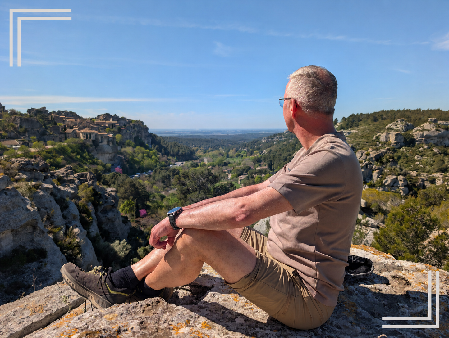 Ein Mann sitzt auf einem Felsen im Freien, blickt auf eine grüne, hügelige Landschaft mit Häusern und Bäumen bei strahlendem Sonnenschein.