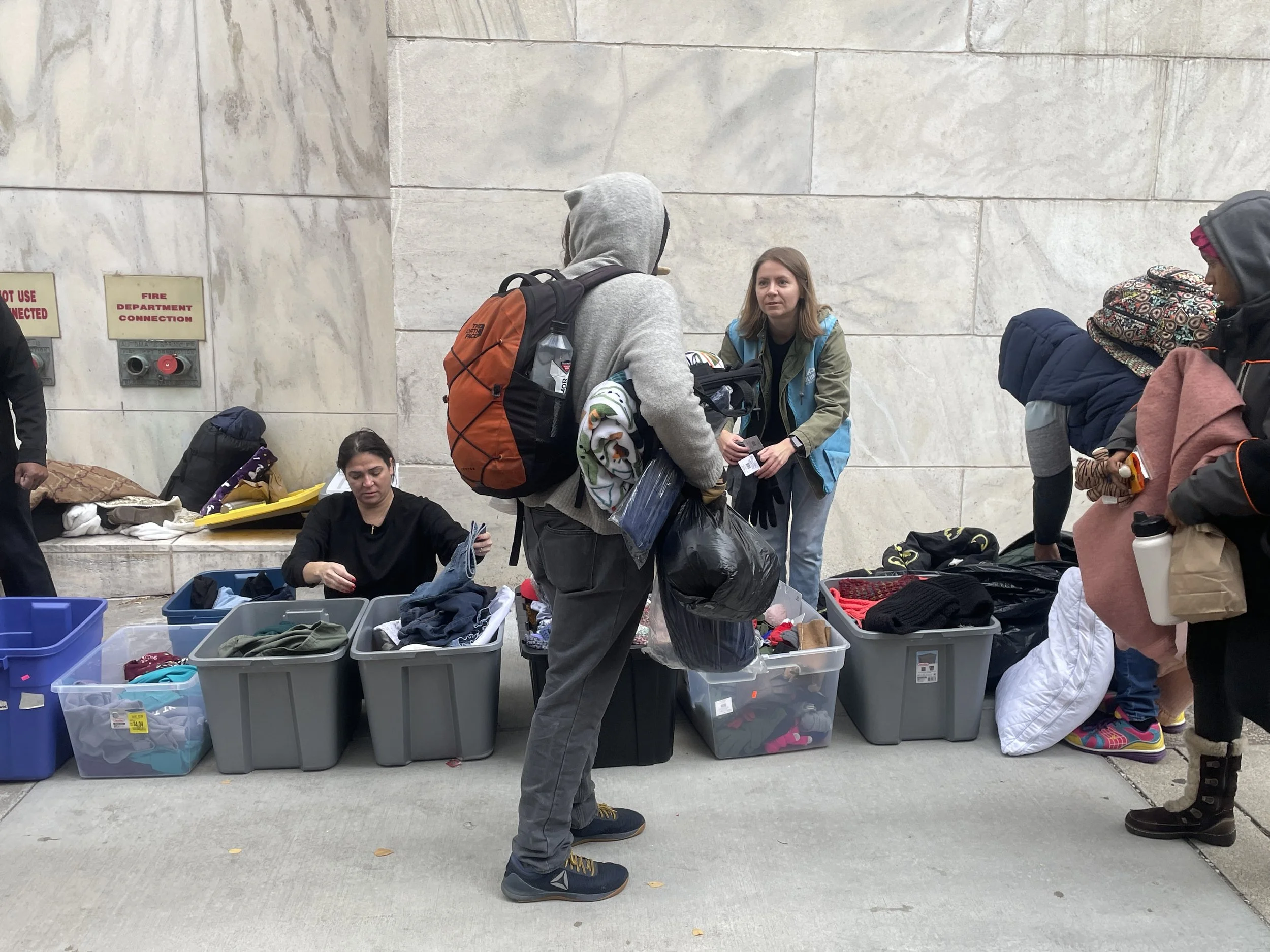 People getting help at a homeless outreach event against a stone wall. Multiple plastic containers filled with clothes are on display, and customers are browsing and conversing.