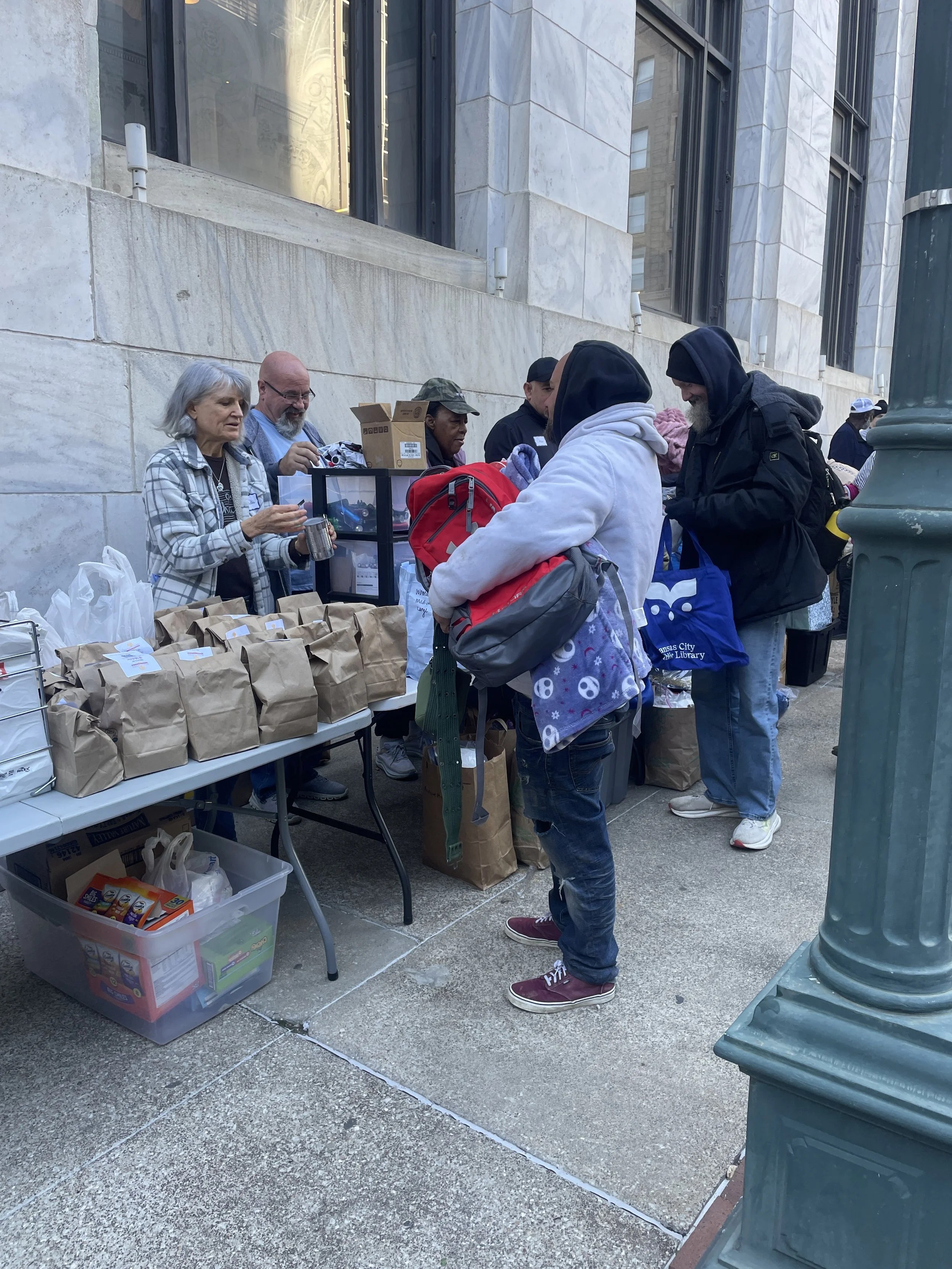 People gather around a table with bags of supplies at a street distribution event in an urban area with tall buildings. Some people are interacting with volunteers behind the table.