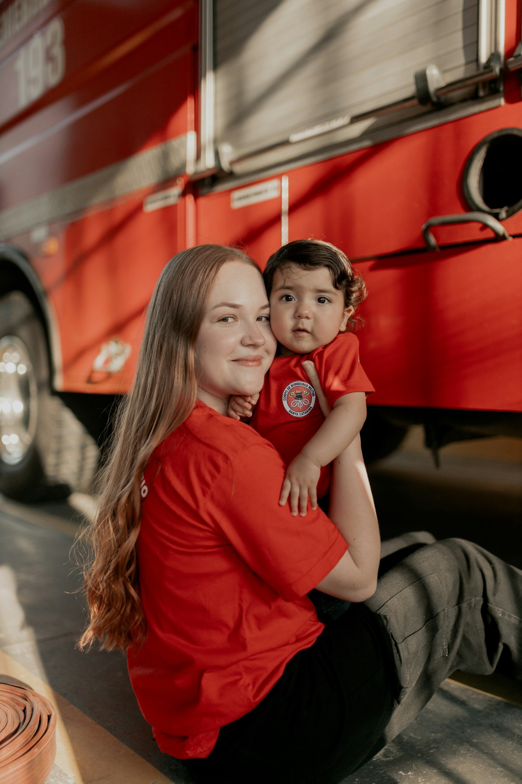 A woman with long red hair and a young boy with curly hair are sitting on the ground in front of a red fire truck, both wearing matching red shirts. The woman is smiling at the camera while holding the boy, who is looking slightly surprised or curious.