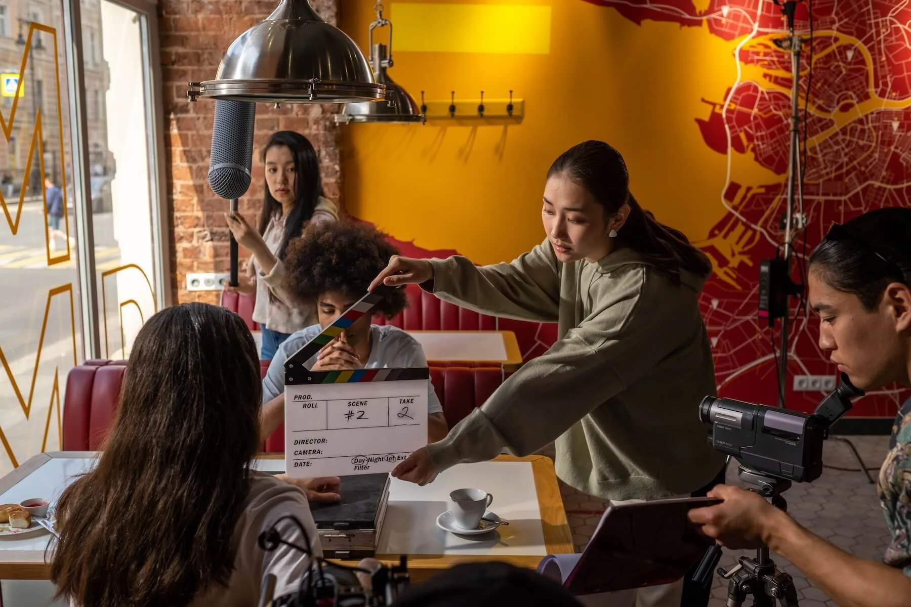 A film crew shooting a scene in a diner, with one crew member holding a clapperboard in front of two seated actors.