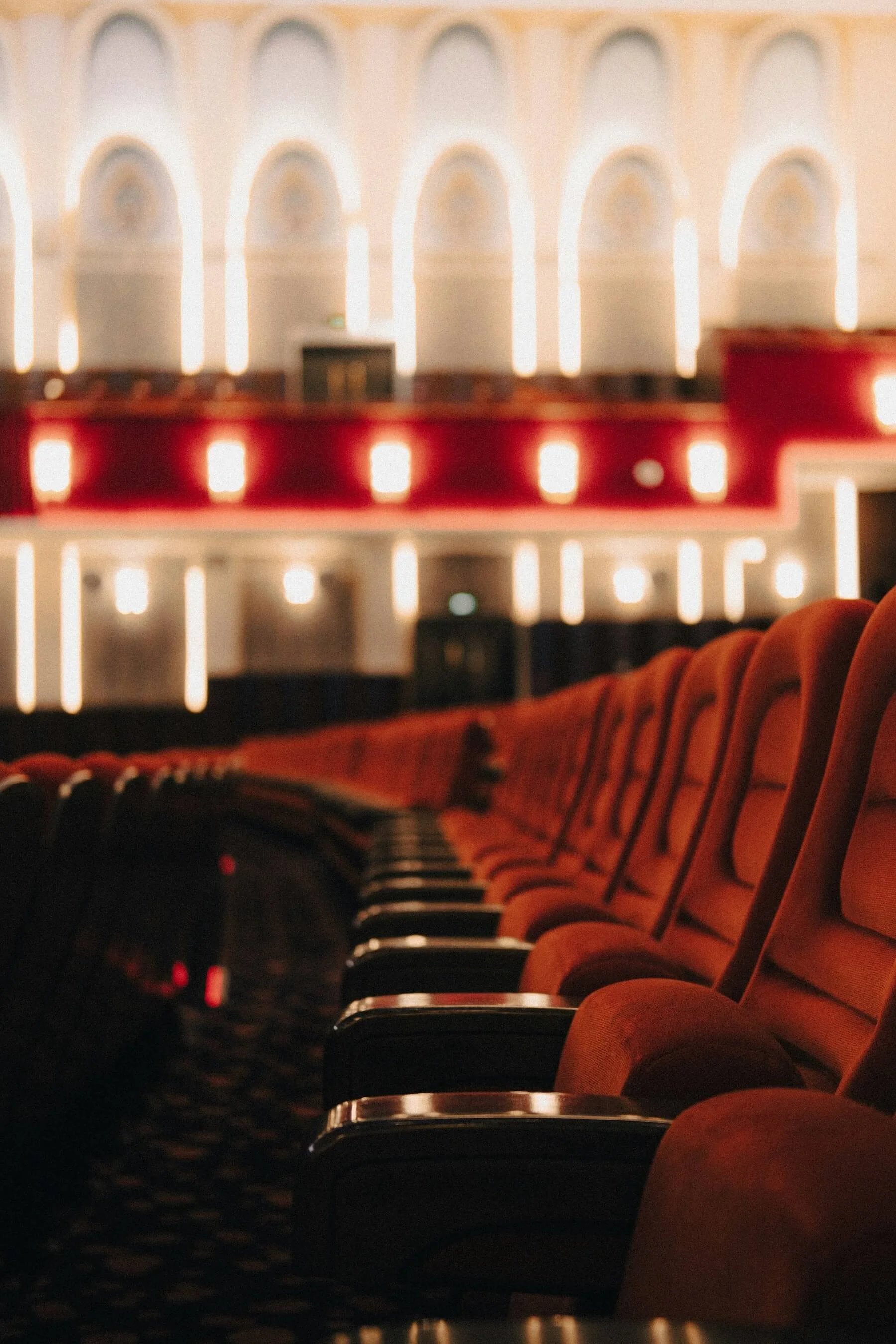 Rows of empty red velvet seats in a historic theater, with a lit balcony blurred in the background.
