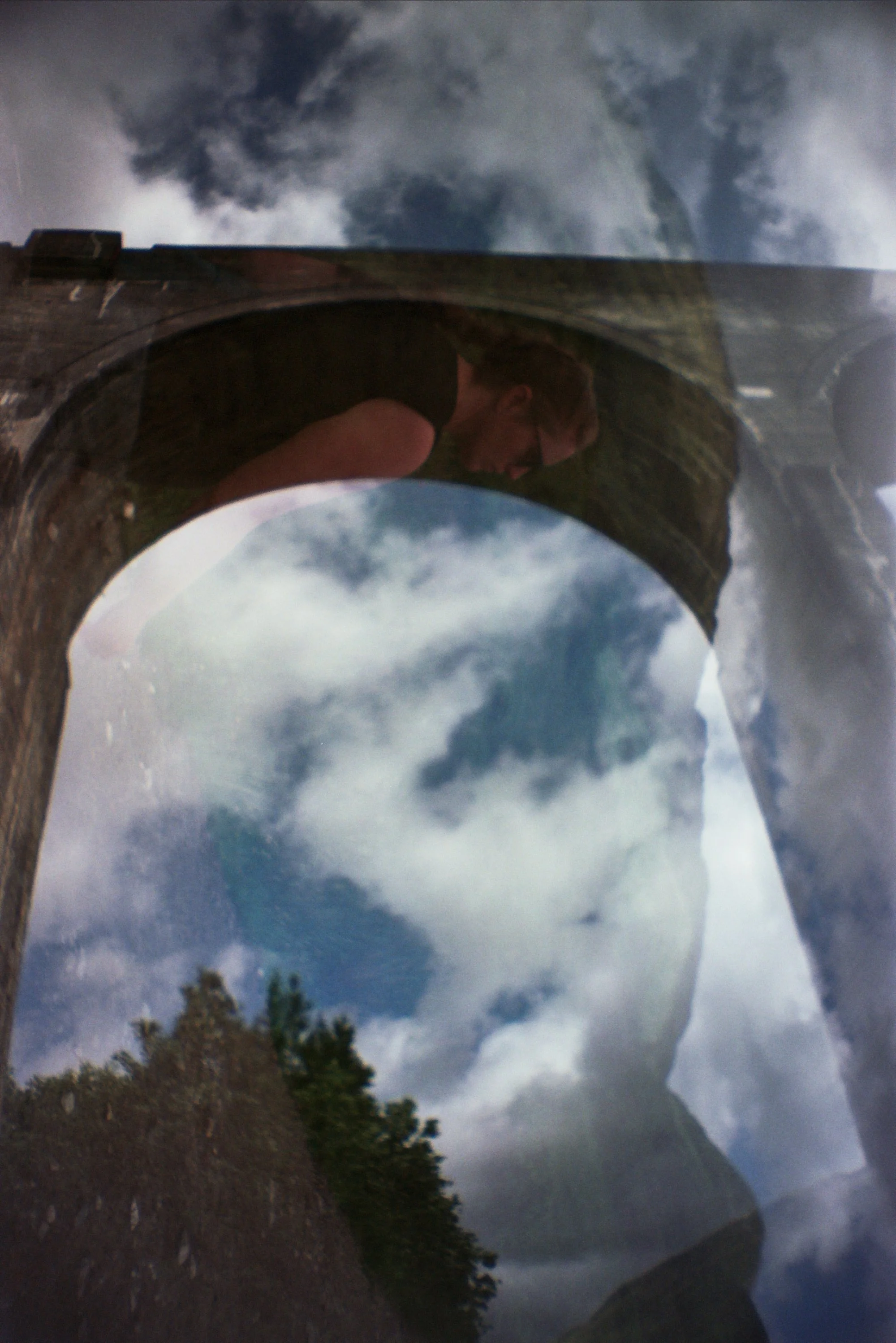 A reflecting surface shows a cloudy sky with dark storm clouds and a person wearing glasses and a black shirt, with trees and a hillside in the background.