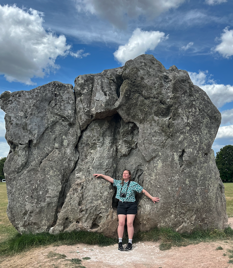 A woman standing with arms outstretched in front of a large, irregularly shaped boulder outdoors under a blue sky with clouds.