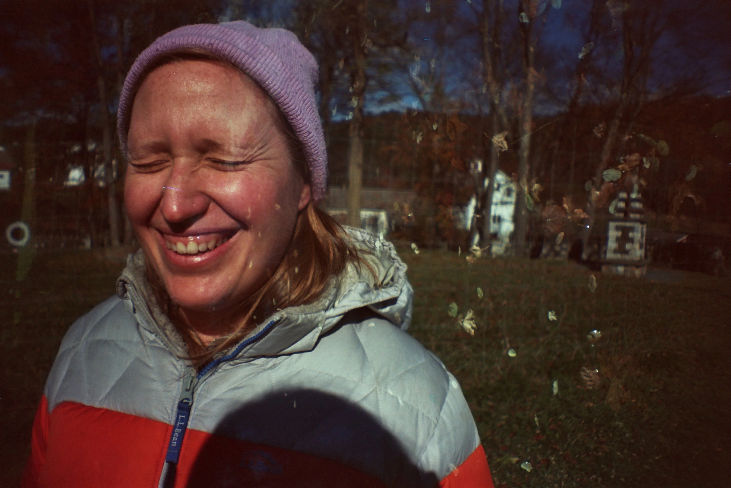A woman with red hair and freckles, wearing a pink knit hat and gray jacket, smiling with her eyes closed outdoors during what appears to be autumn, with trees and houses in the background.