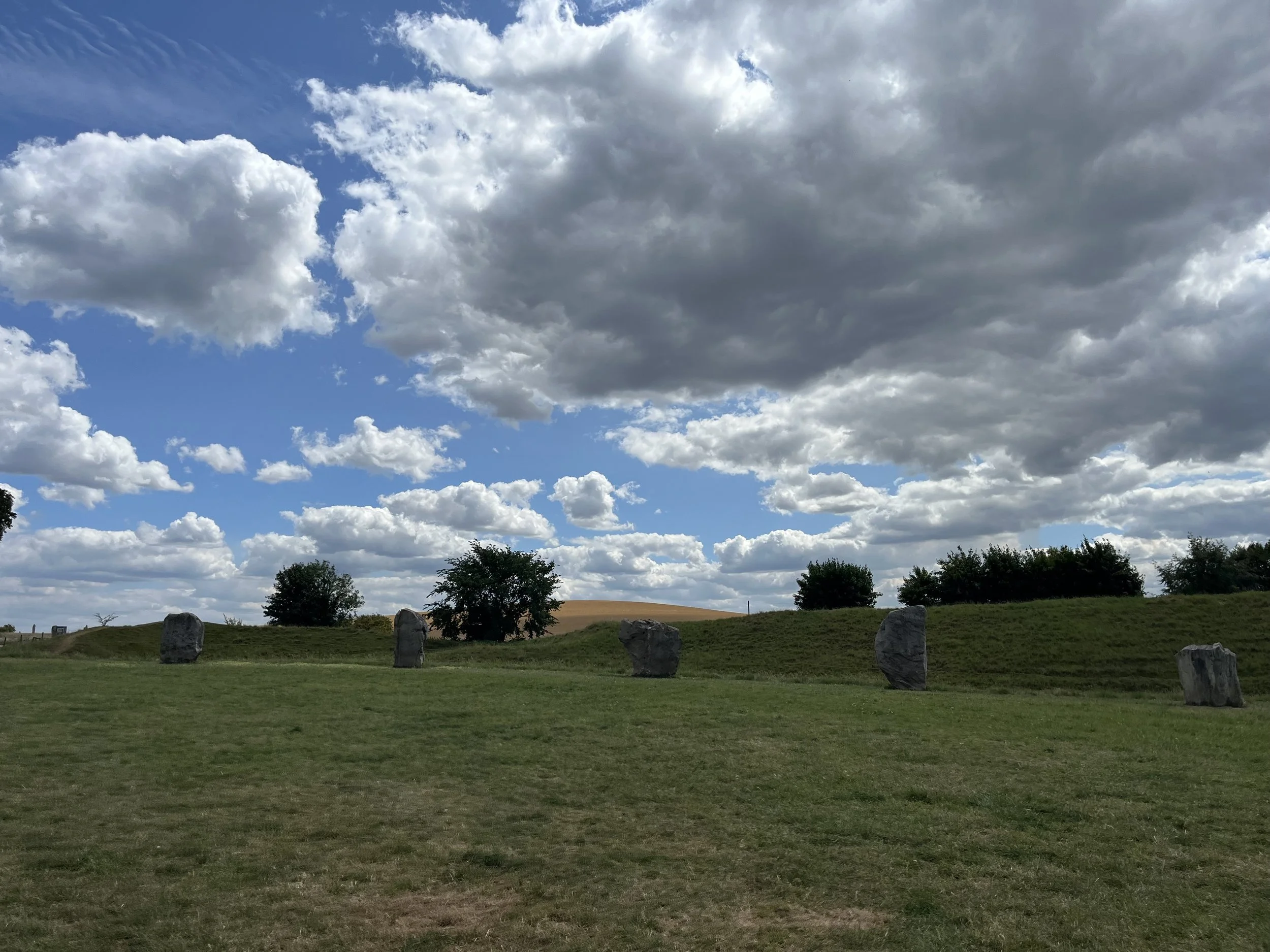 A landscape scene with a grassy foreground, a few scattered trees, stone markers, a hill in the background, and a partly cloudy sky overhead.