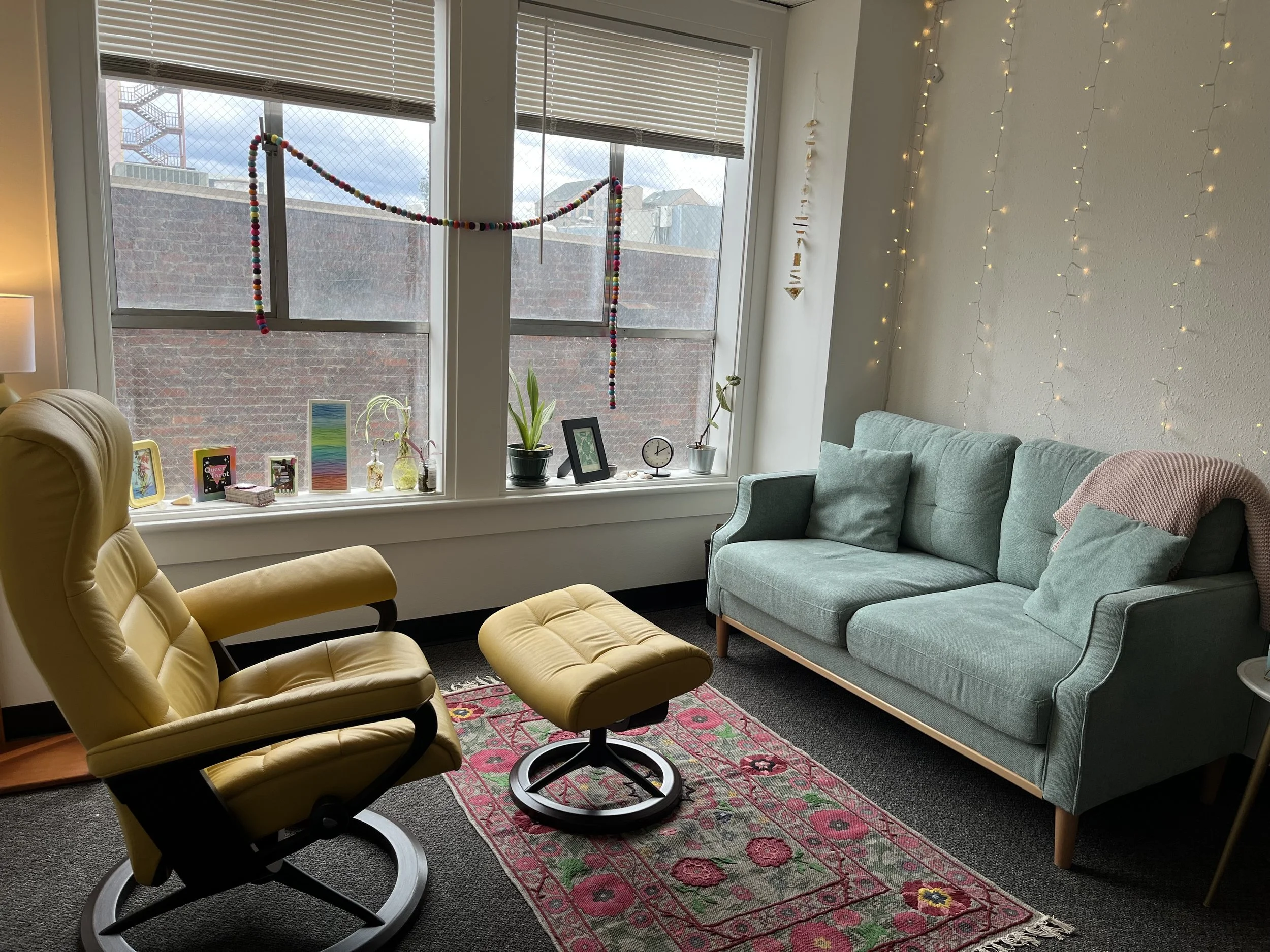 A living room with a large window, yellow armchair, mint green sofa, and a pink area rug. Decor includes string lights on the wall, small plants, framed pictures, and a colorful garland hanging in the window.
