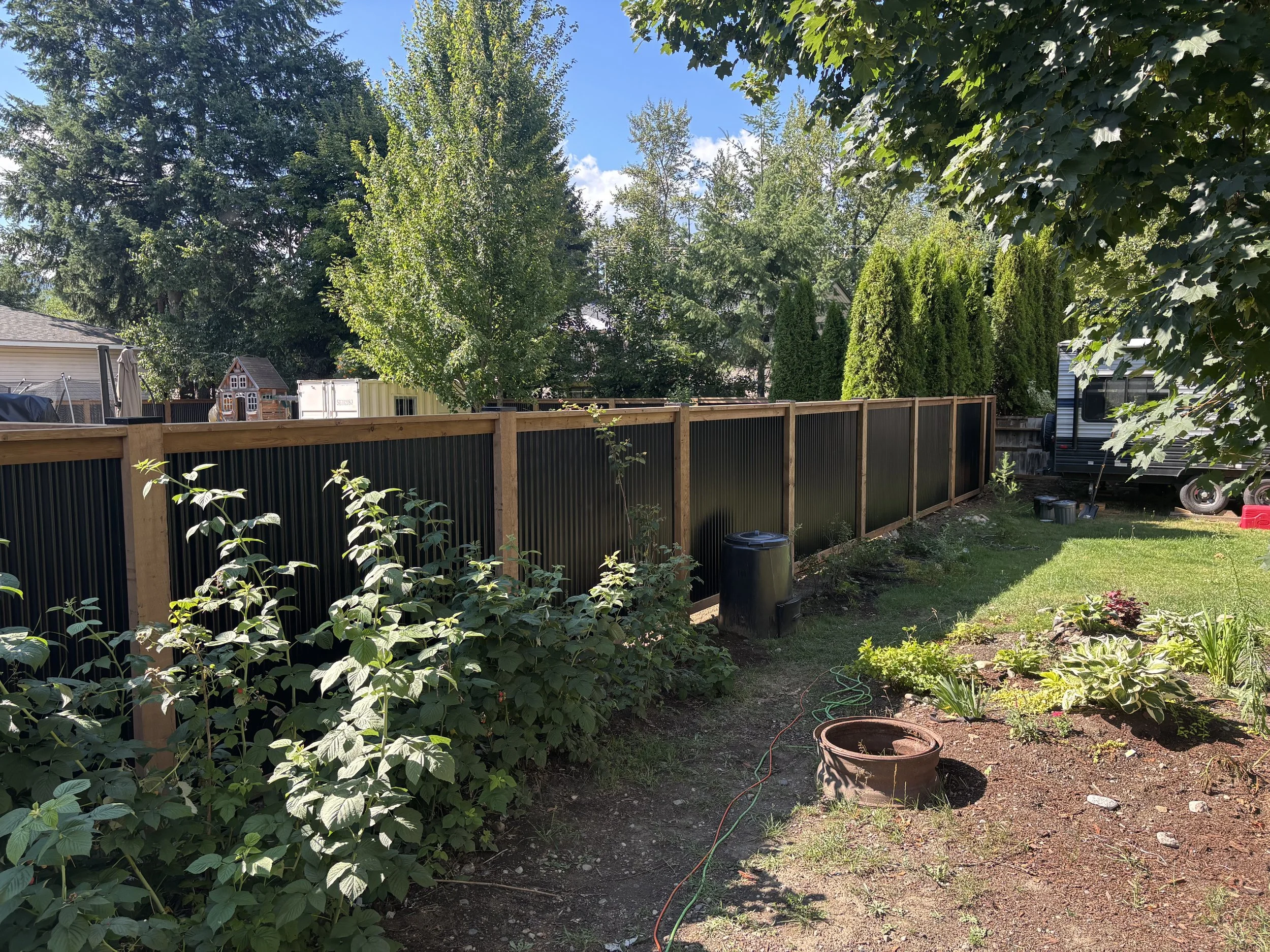 A backyard garden with a wooden fence, various plants, a small playhouse, and tall trees under a blue sky.