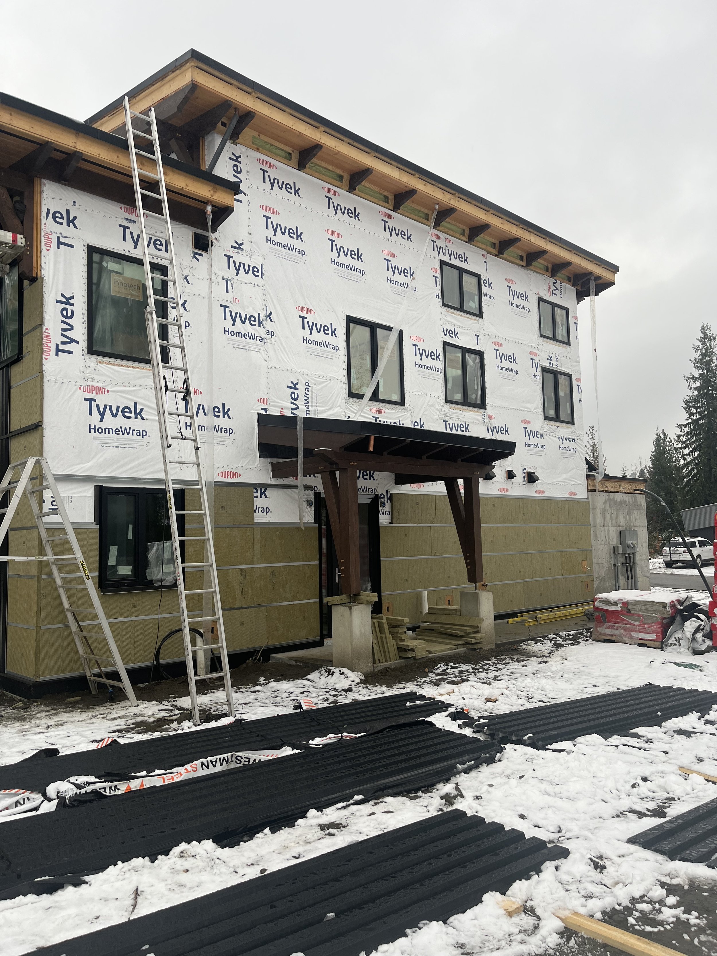 A building under construction with partially installed siding, a ladder leaning against it, and construction materials scattered on snow-covered ground.