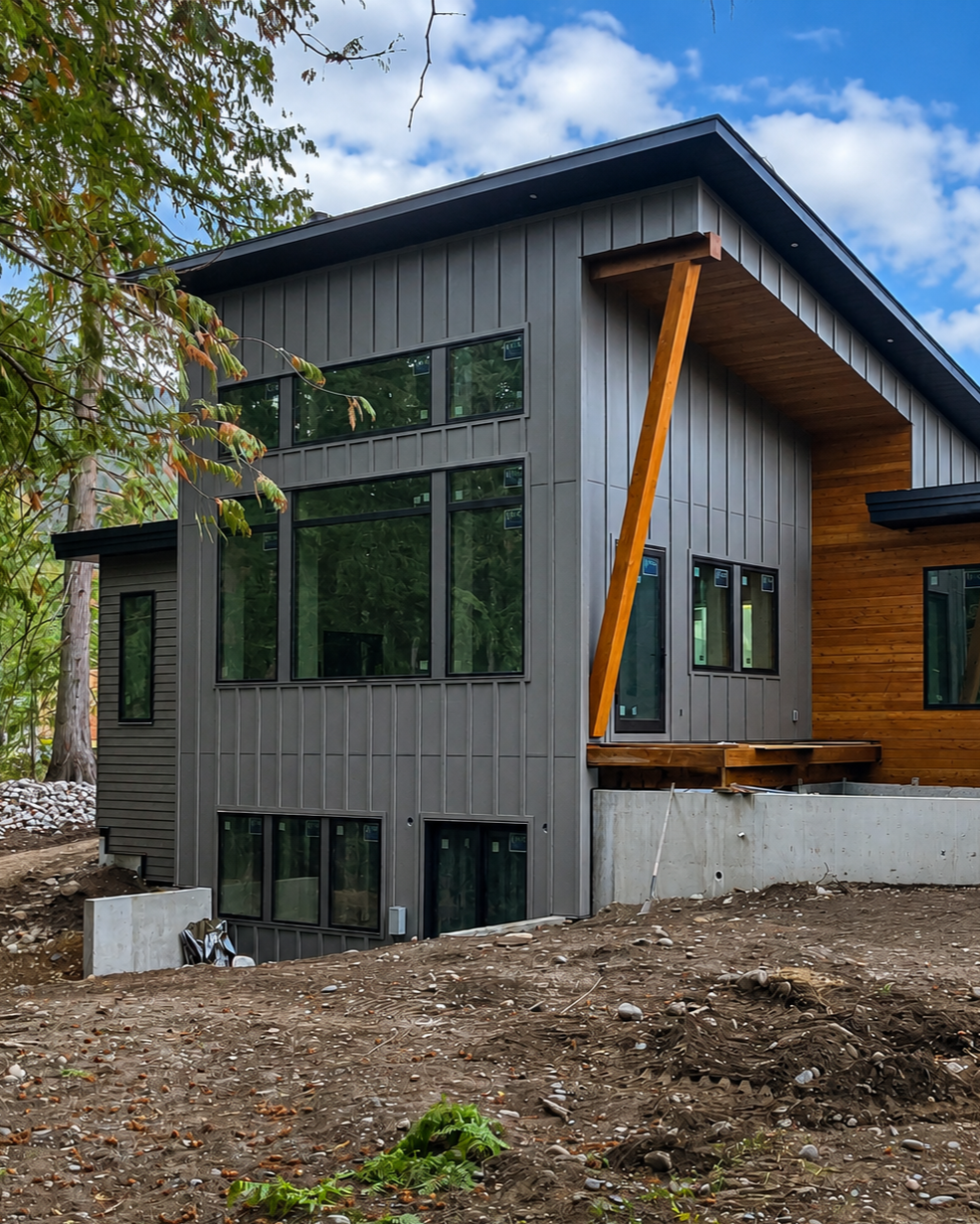 Modern multi-story house with gray and brown exterior, large windows, and an overhanging roof, located in a natural setting with trees and a partly cloudy sky.