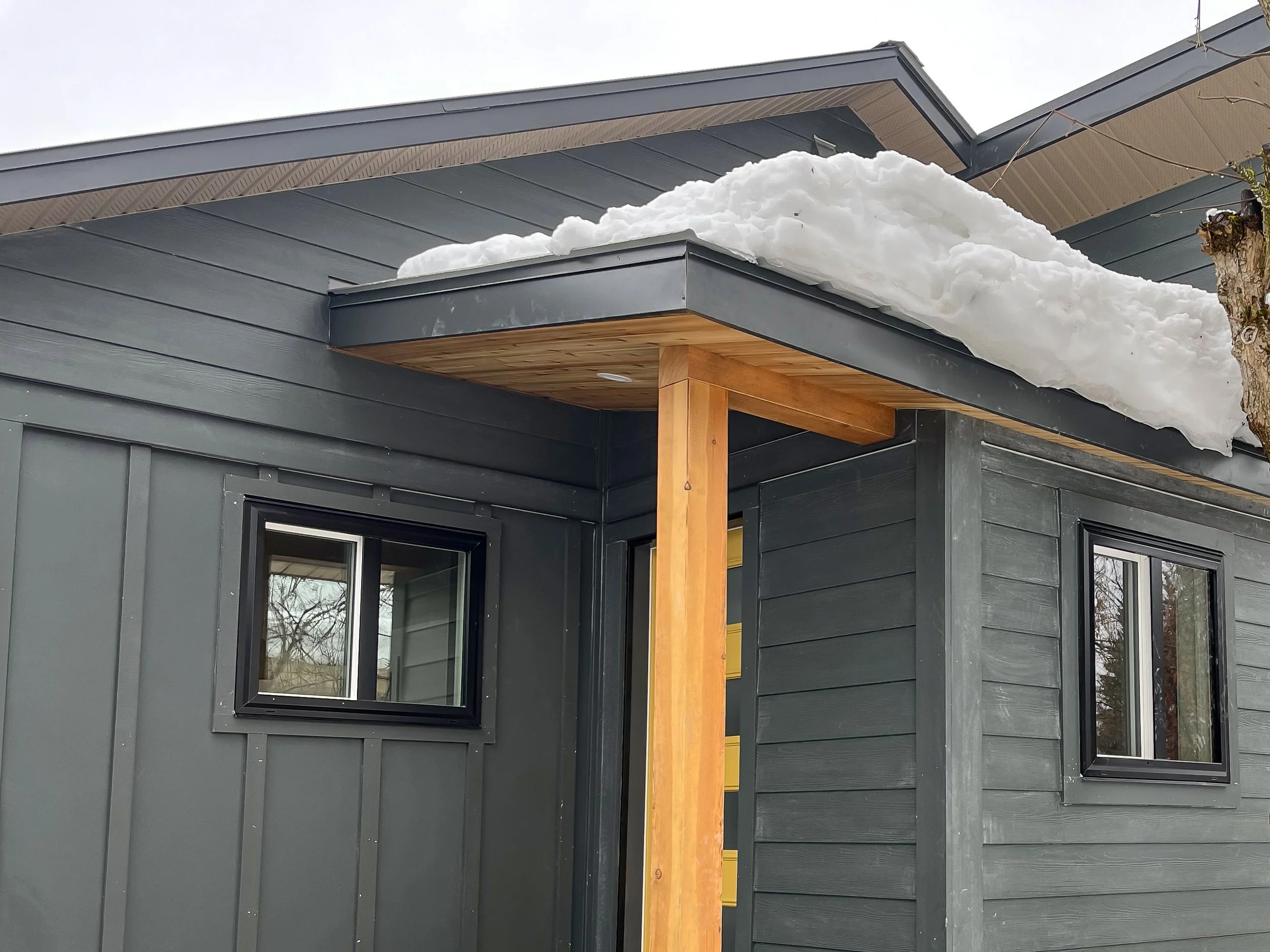 A house with a dark gray exterior, black-framed windows, and a modern architectural design. There is a wooden support beam on the front porch, and snow is resting on the roof and the porch roof.
