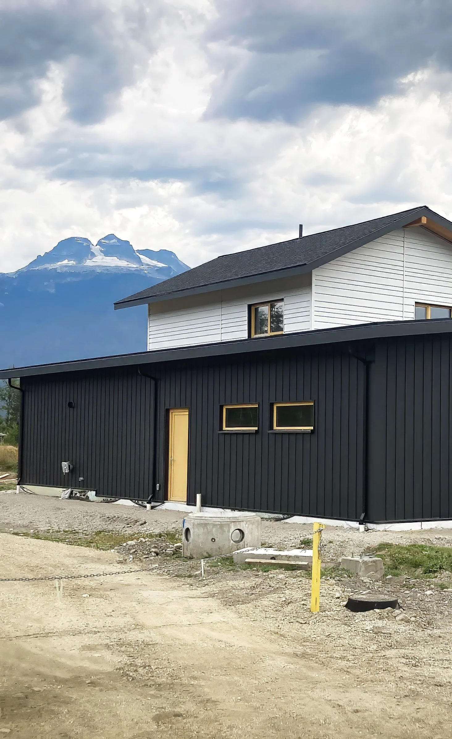 Photo of a house under construction, with black and white siding, a yellow door, and a mountain range with snow on the peaks in the background. The ground is dirt with construction materials around.