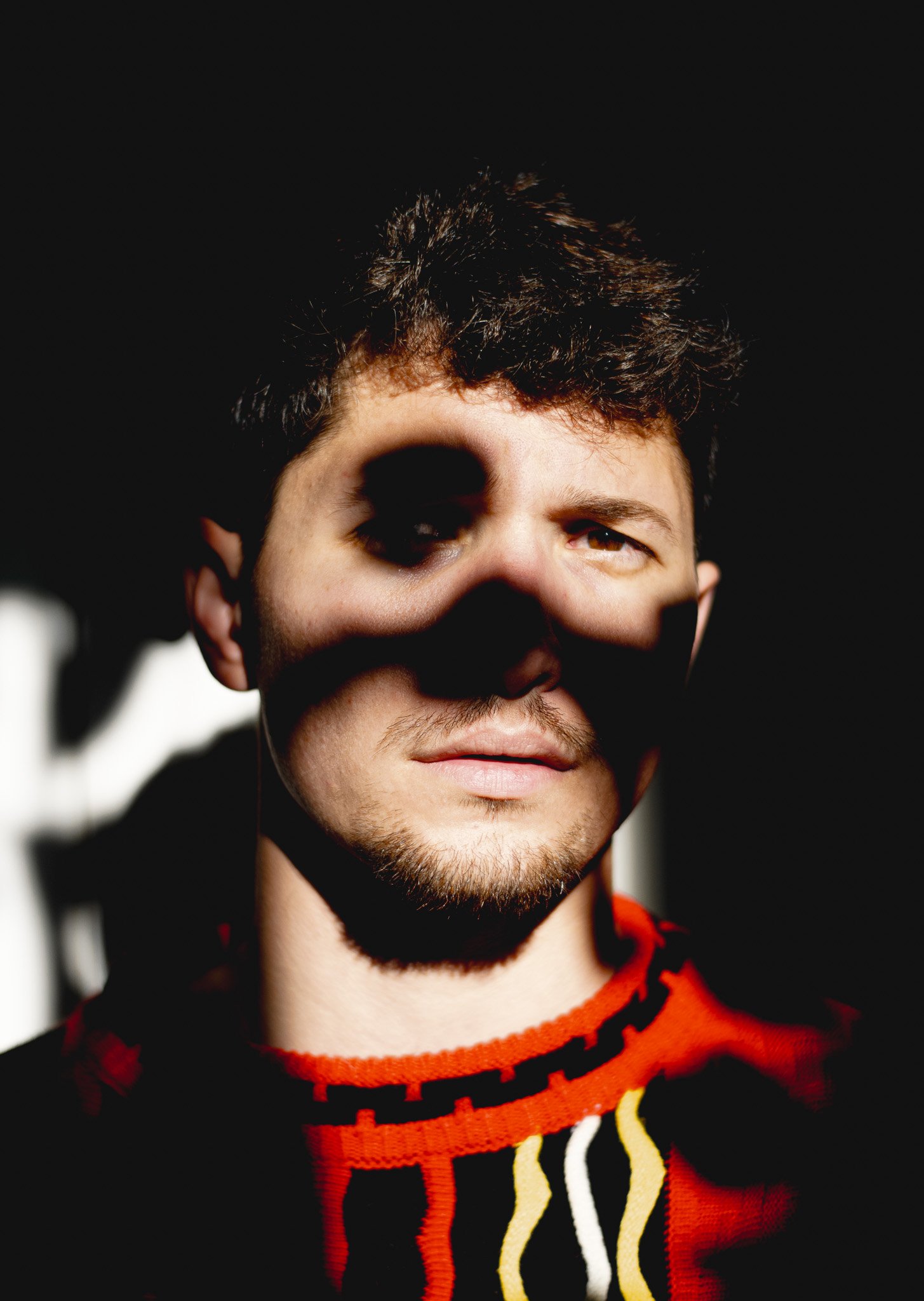 Close-up of a young man with dark hair and a goatee, wearing a red and black patterned sweater, against a dark background.