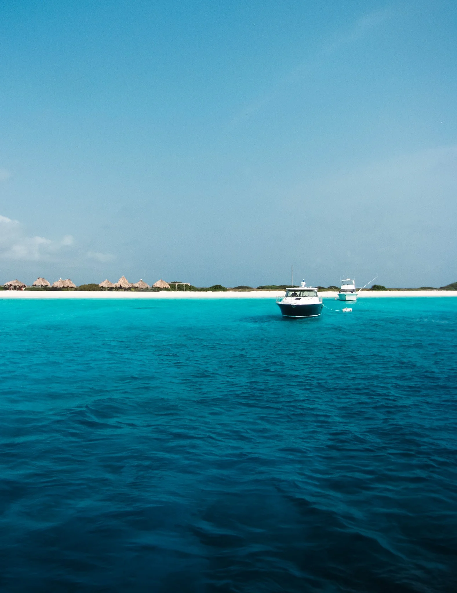 Calm turquoise ocean water with two boats anchored near the shore, under a light blue sky with some clouds, a sandy beach with straw umbrellas and greenery in the background.