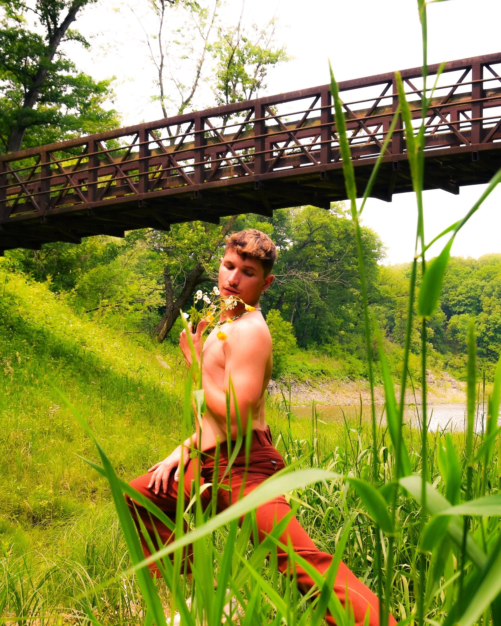 A young man sitting in a grassy area near a river, holding a bouquet of white flowers, with a wooden bridge and green trees in the background.
