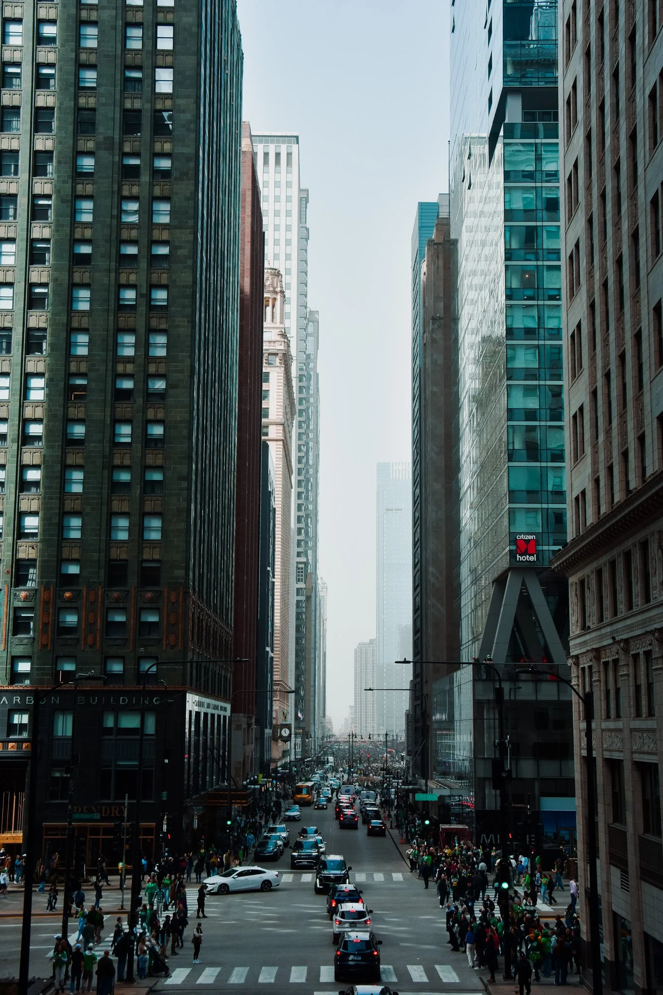 A busy city street scene with cars, taxis, and pedestrians at a crosswalk, flanked by tall skyscrapers with glass and brick facades.
