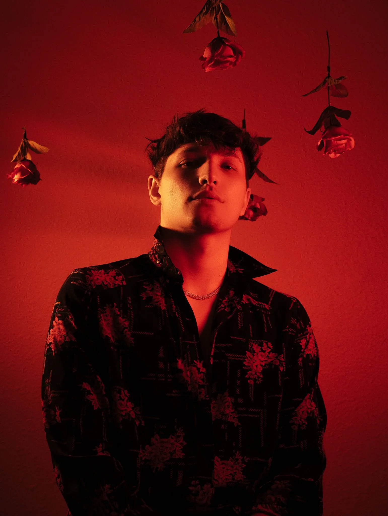 A young man with short wavy hair and light skin, wearing a black floral jacket and silver necklace, standing against a red background with hanging dried roses.