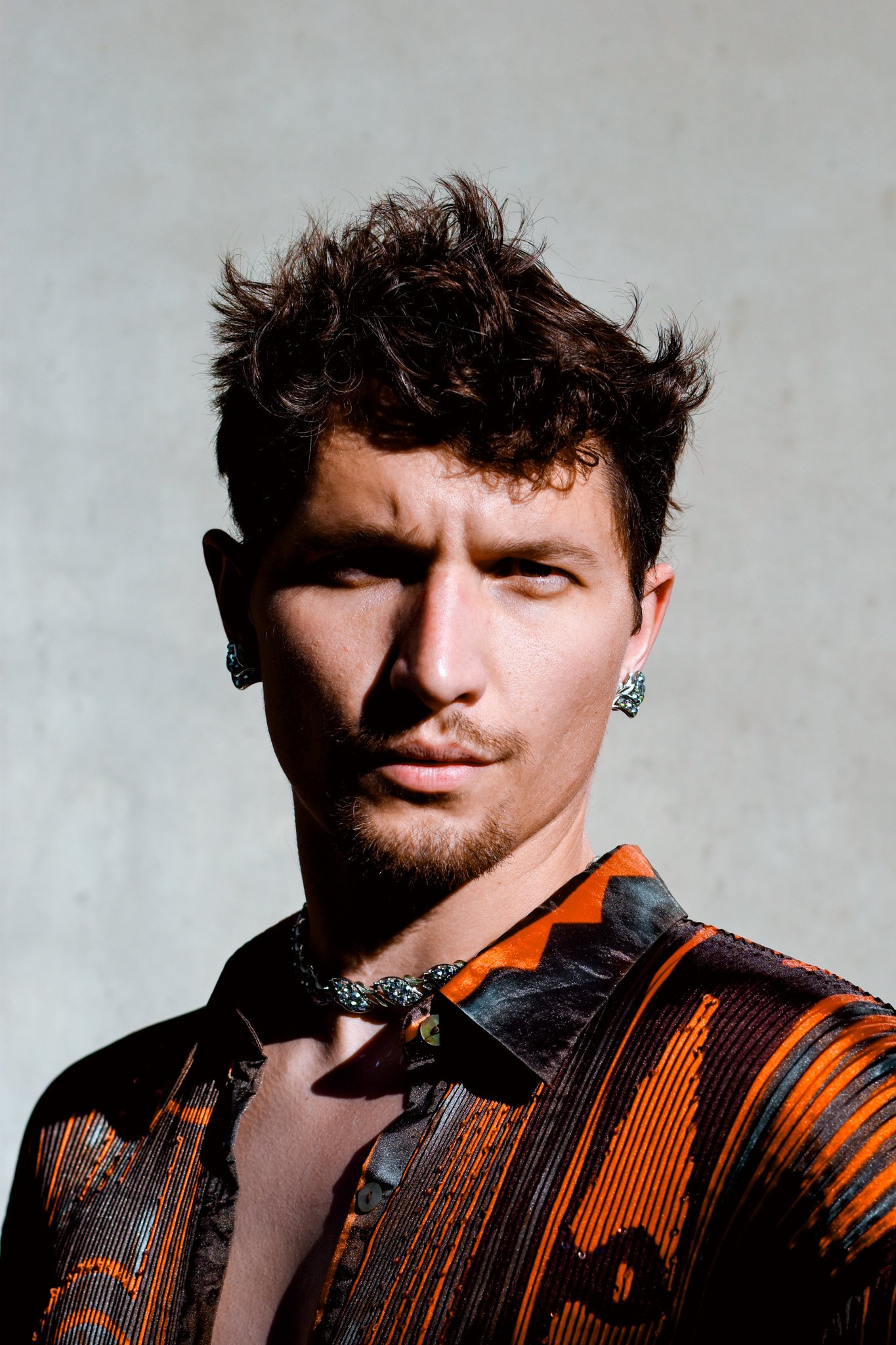 Close-up of a person with short, curly dark hair, wearing earrings, a patterned jacket, and a statement necklace, with sunlight casting shadows on their face, against a plain background.