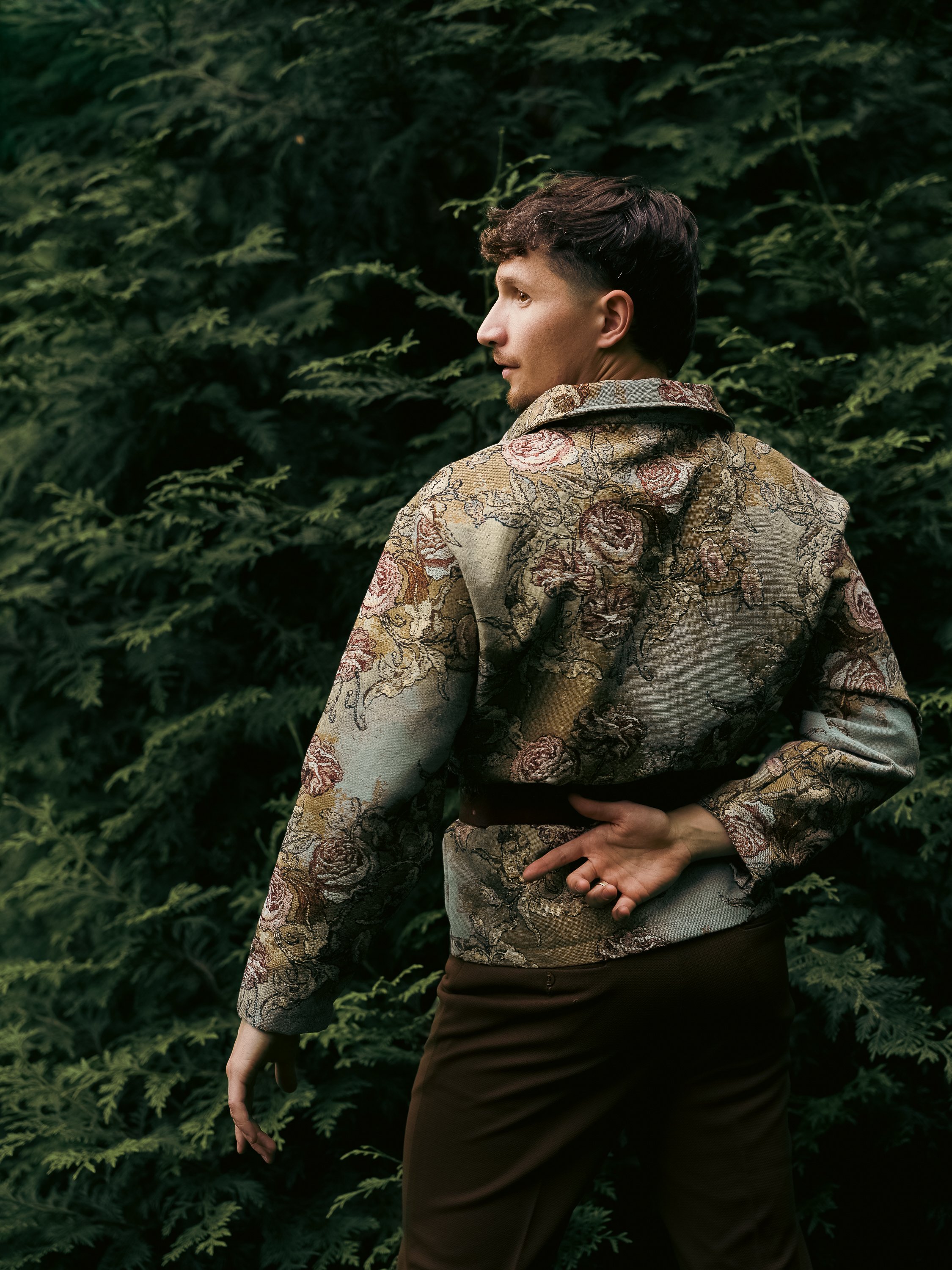A person standing outdoors in front of lush green foliage, wearing a floral patterned jacket and dark pants, with one hand resting on their hip and the other hanging down.