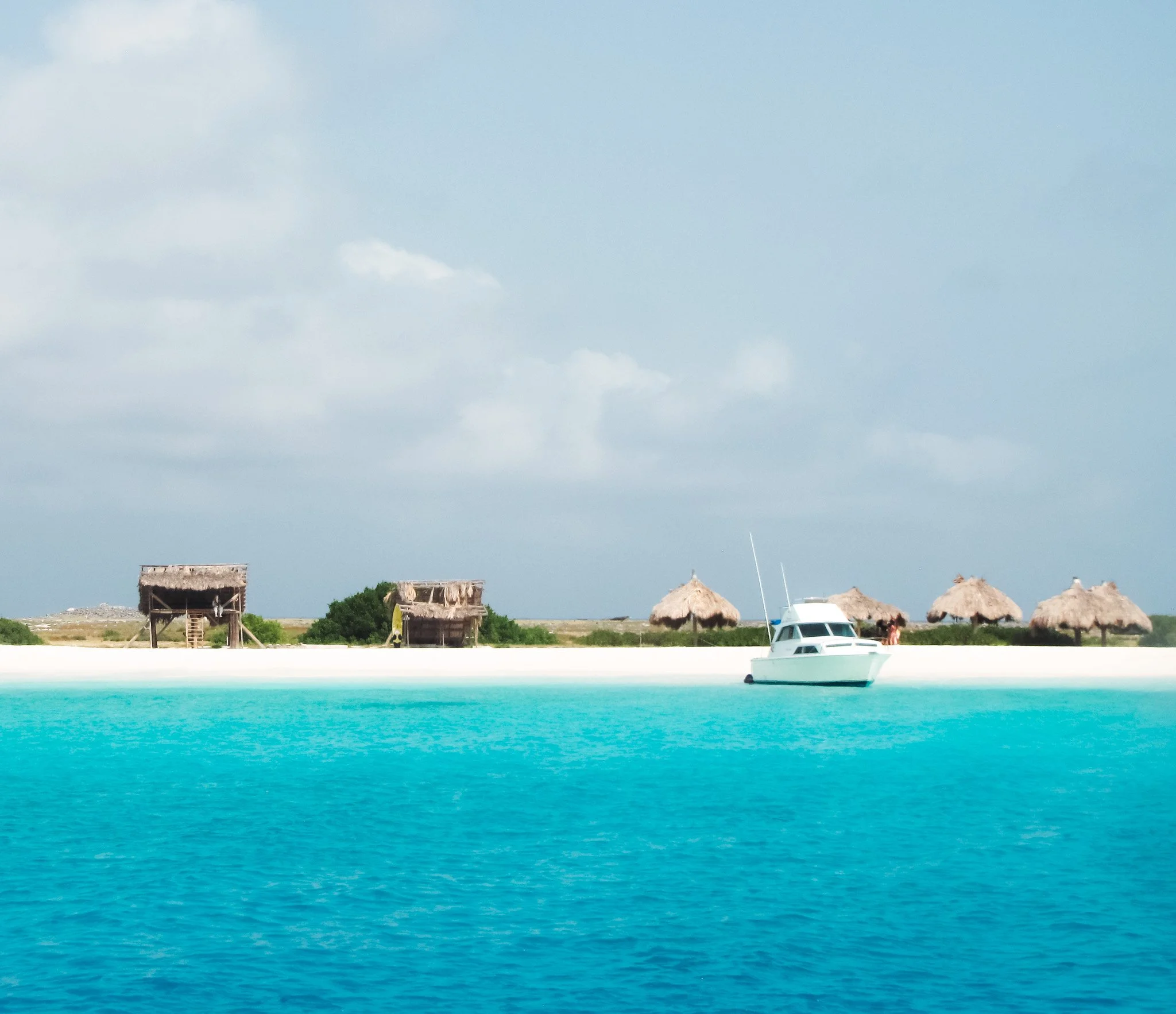 Calm tropical waters with a white boat, beach huts, and thatched roofs along the shore, under a cloudy sky.