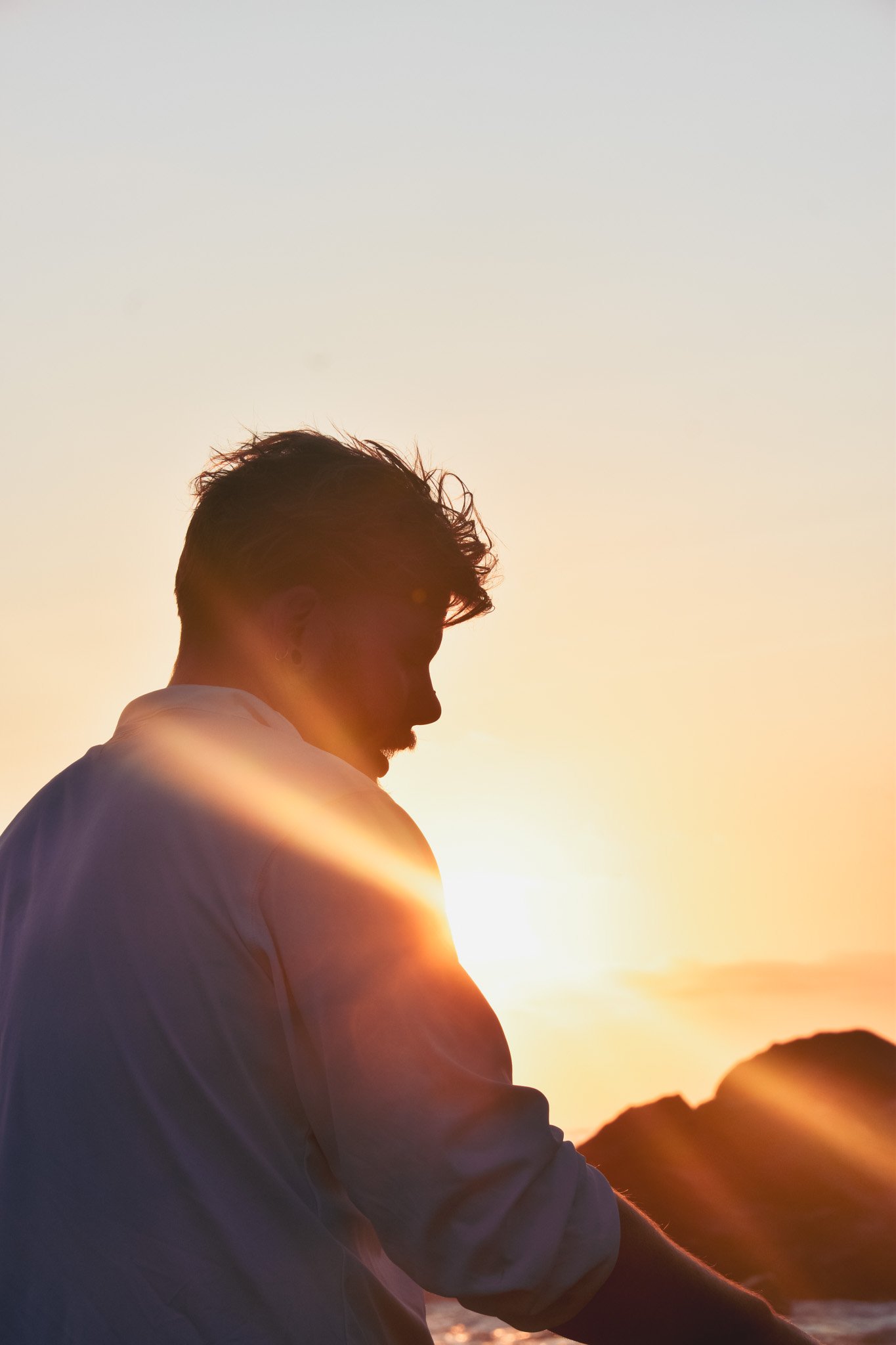 A person with curly hair wearing a white shirt, sitting outdoors at sunset with mountains in the background.