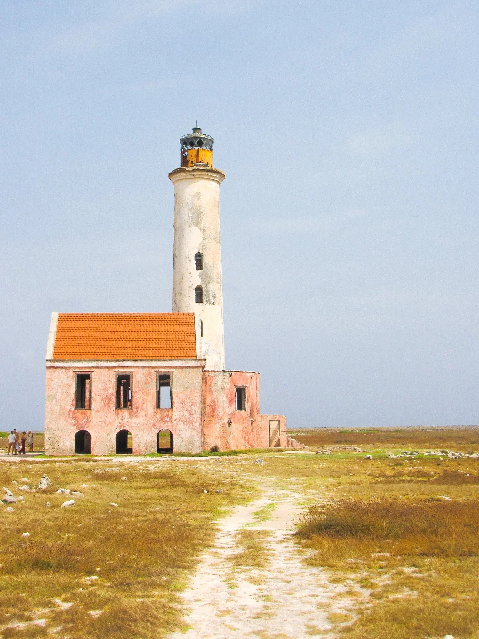 A lighthouse with a weathered white tower and a black and yellow lantern room, attached to a pinkish building with small windows and arched doorways, situated in an open field with a dirt path leading towards it, under a clear blue sky.