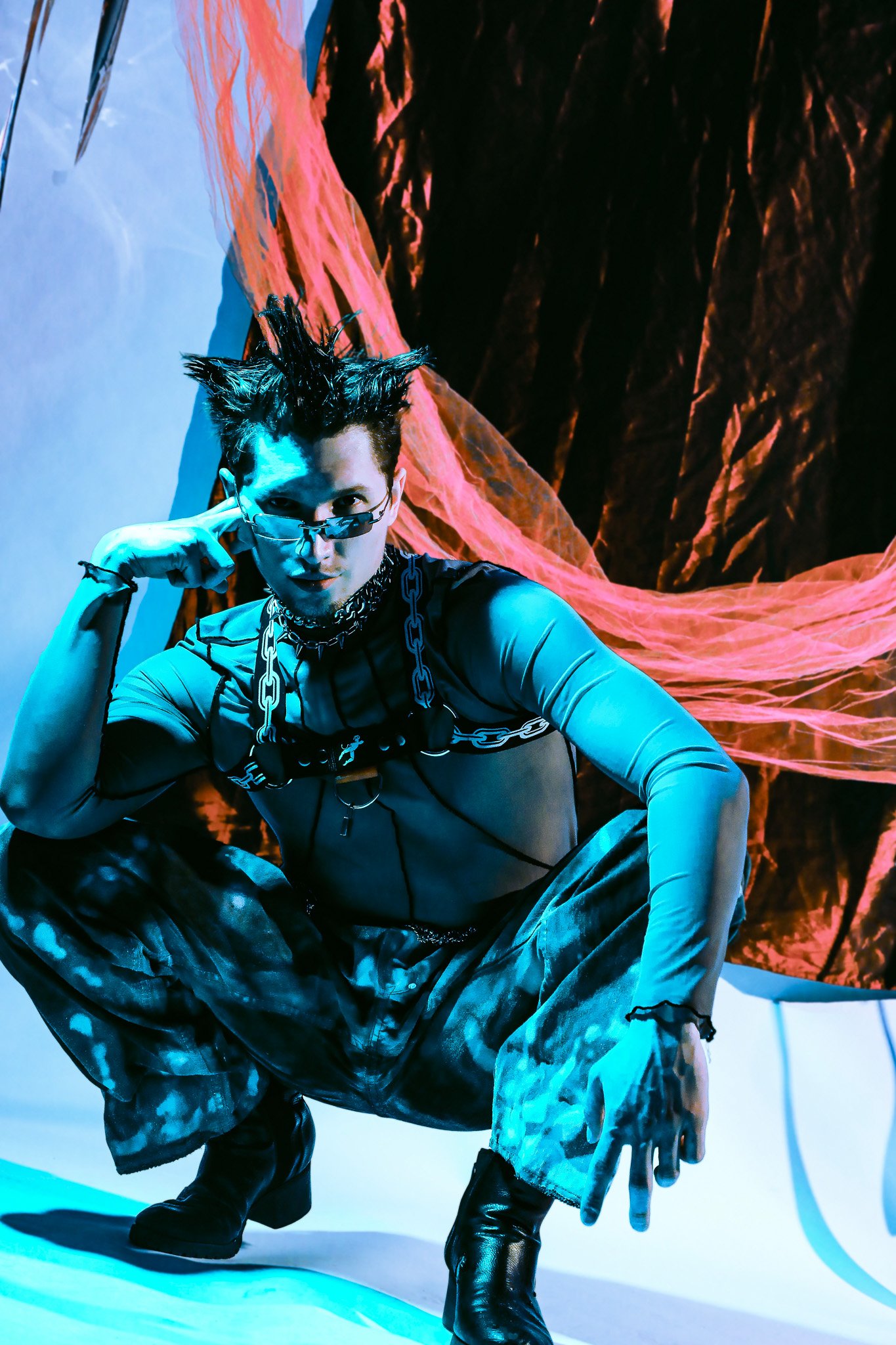 A person with spiked hair, wearing sunglasses, a chain necklace, and metallic clothing, crouches in a futuristic setting with vibrant blue and orange lighting.