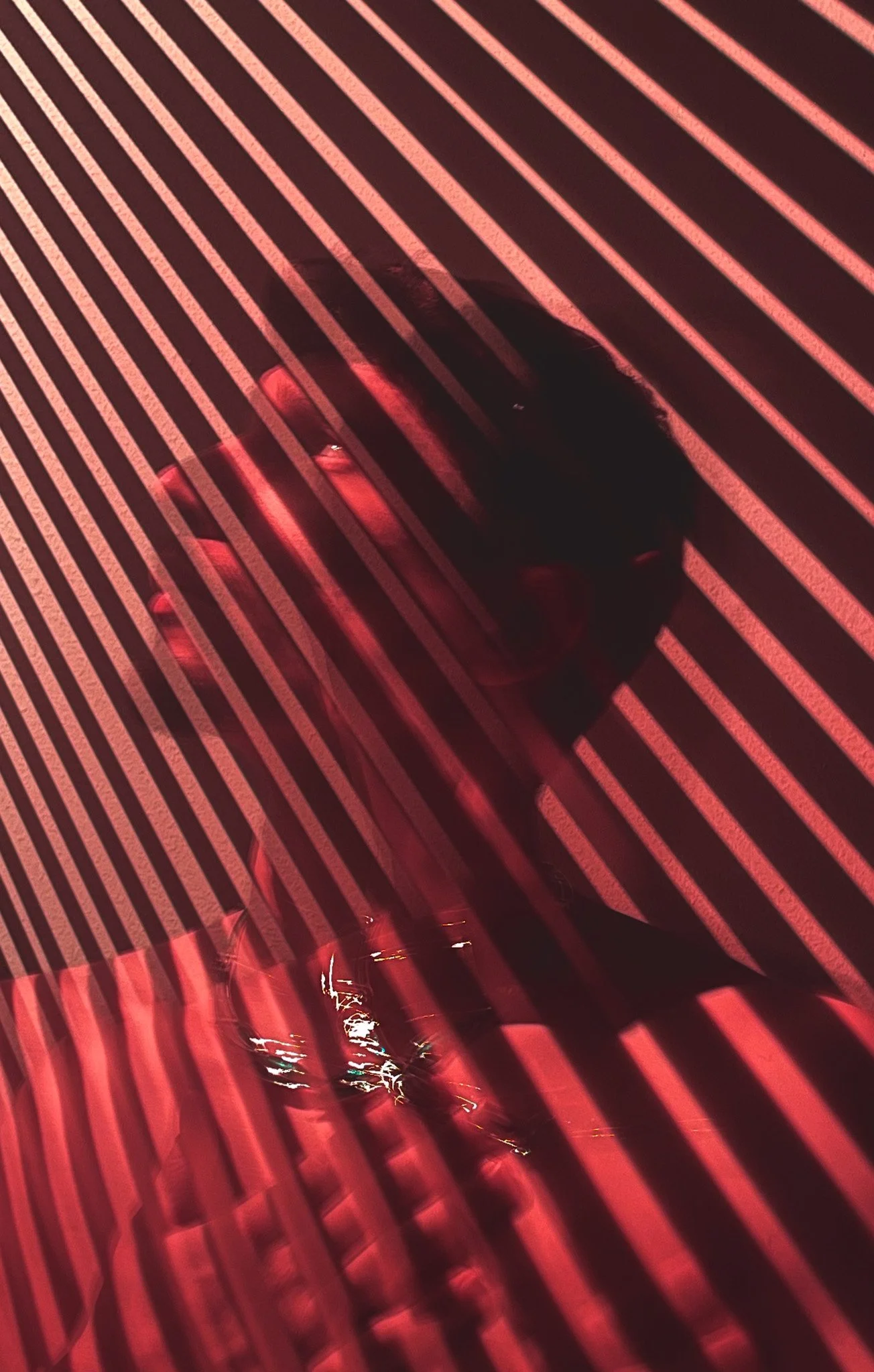 Person in dark clothing and sneakers walking on a street illuminated with red neon lights, seen through a pattern of red and white stripes.