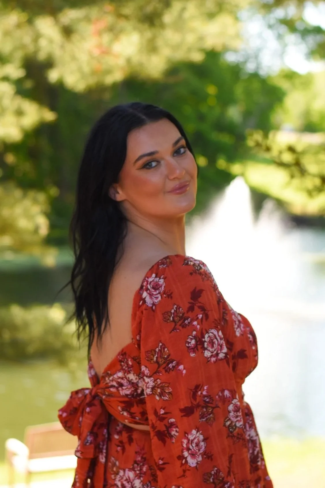 A woman with dark hair, wearing a reddish-orange floral dress, standing outdoors near a body of water and trees in the background.