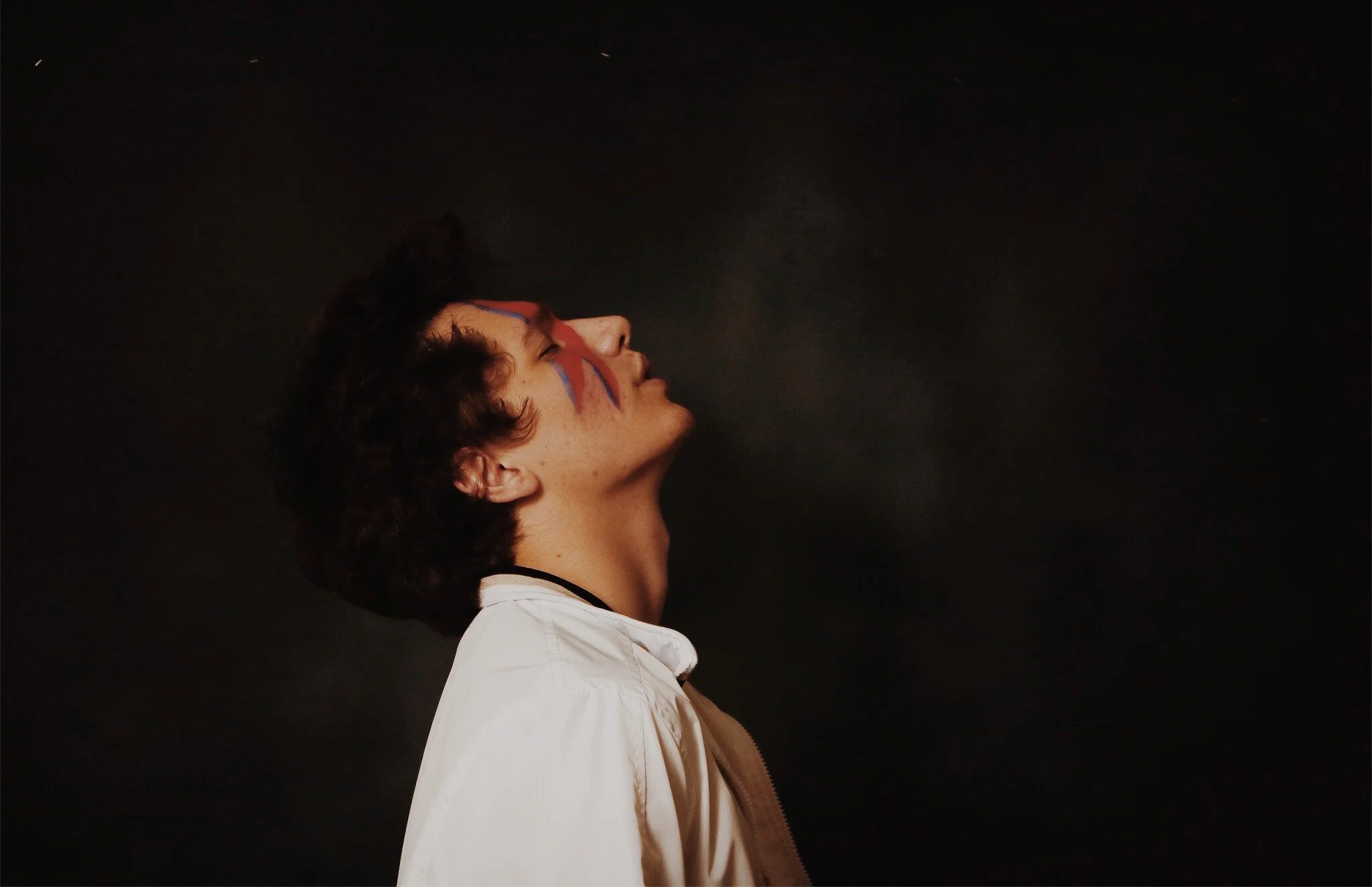 Side profile of a young man with face paint, wearing a white shirt, against a dark background.