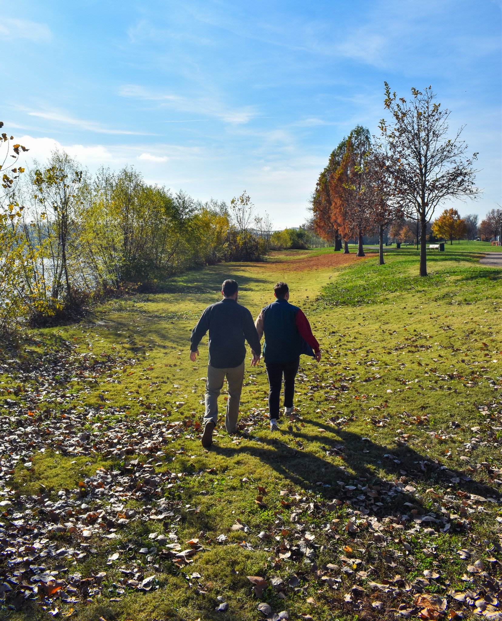Two men walking in a park on a sunny autumn day, with trees and fallen leaves around.