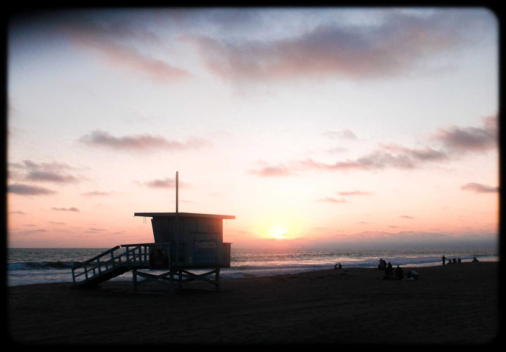 A beach scene during sunset with the sky partly cloudy and a lifeguard tower in the foreground, as people relax by the water.