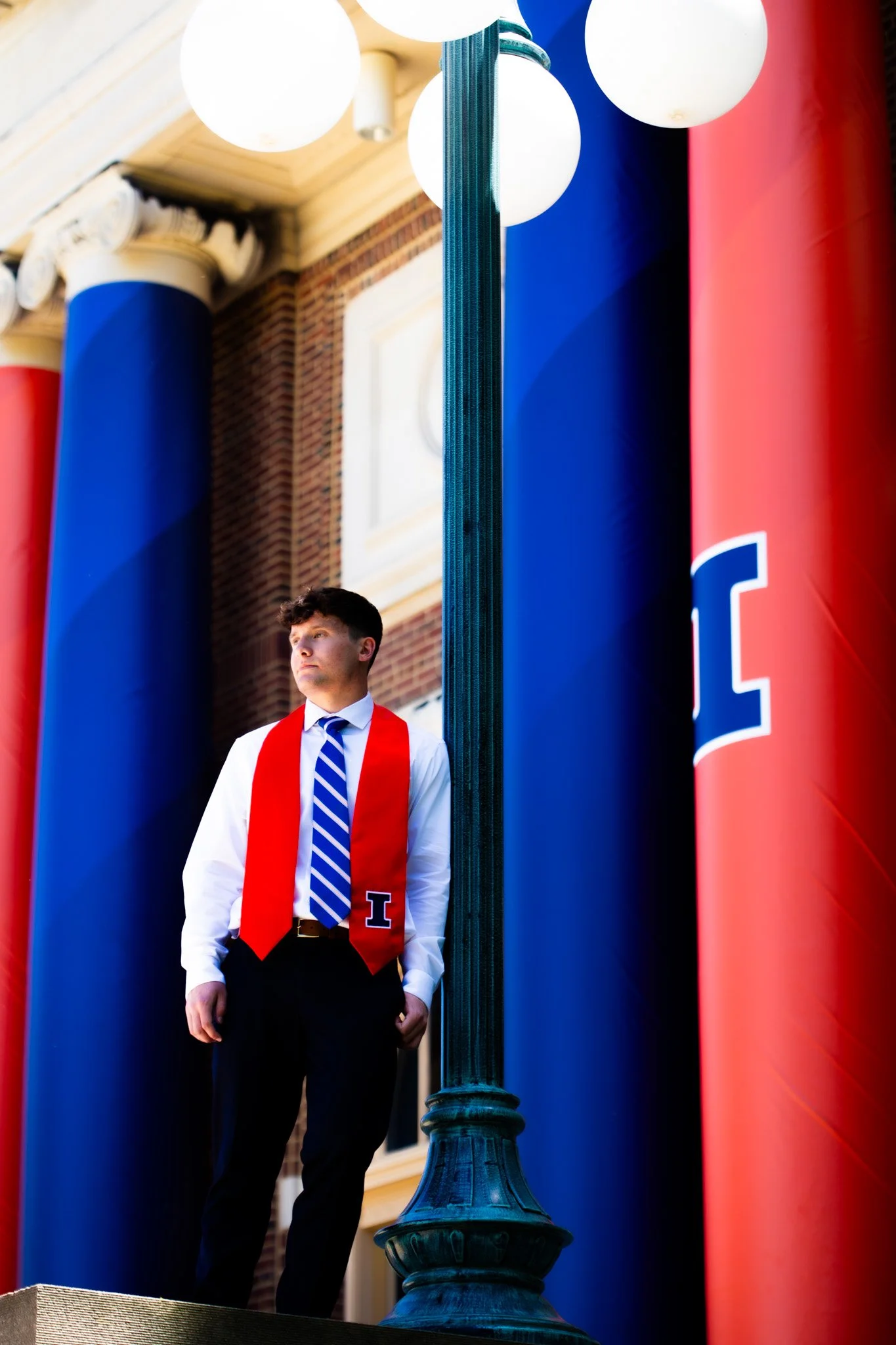 A young man in formal attire, wearing a white shirt, a striped blue and white tie, and a red sash with the letter 'I', stands on a sidewalk. There are large inflatable columns colored in red, blue, and partially visible to the sides, with a lamppost 