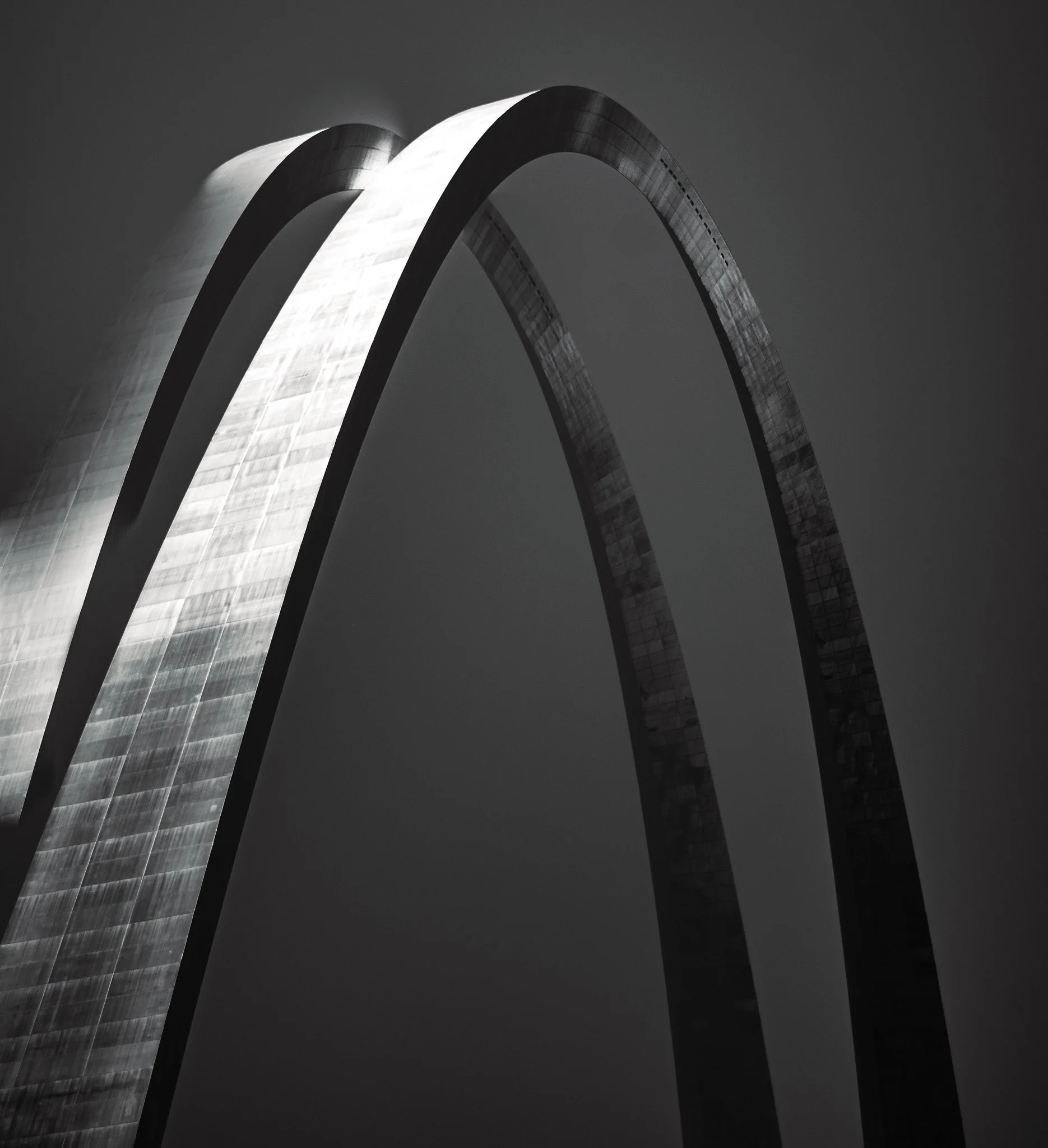 A black and white photo of the Gateway Arch in St. Louis, Missouri, showing two large, curved stainless-steel arches against a dark sky.