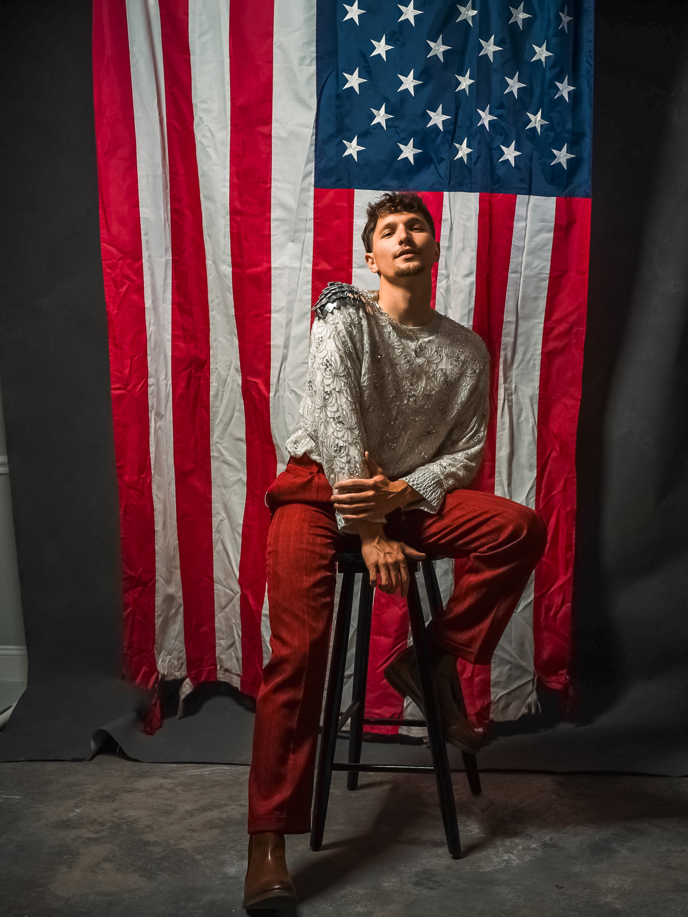 A young man with curly hair, a goatee, wearing a shiny patterned shirt, red pants, and brown boots, sitting on a stool in front of an American flag backdrop with black sides.