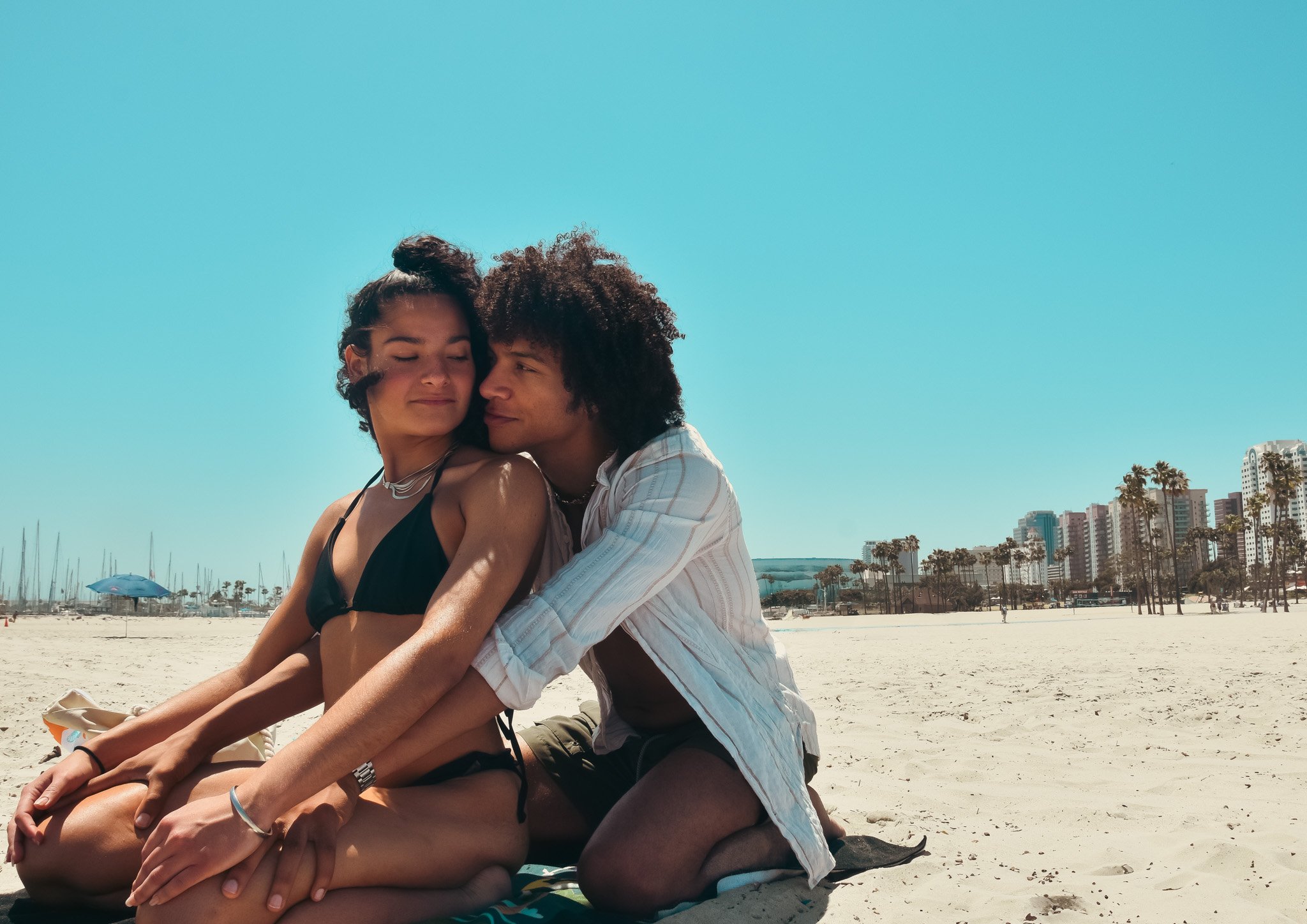 A young couple sitting on the beach with closed eyes, embracing each other. There are high-rise buildings and palm trees in the background under a clear blue sky.