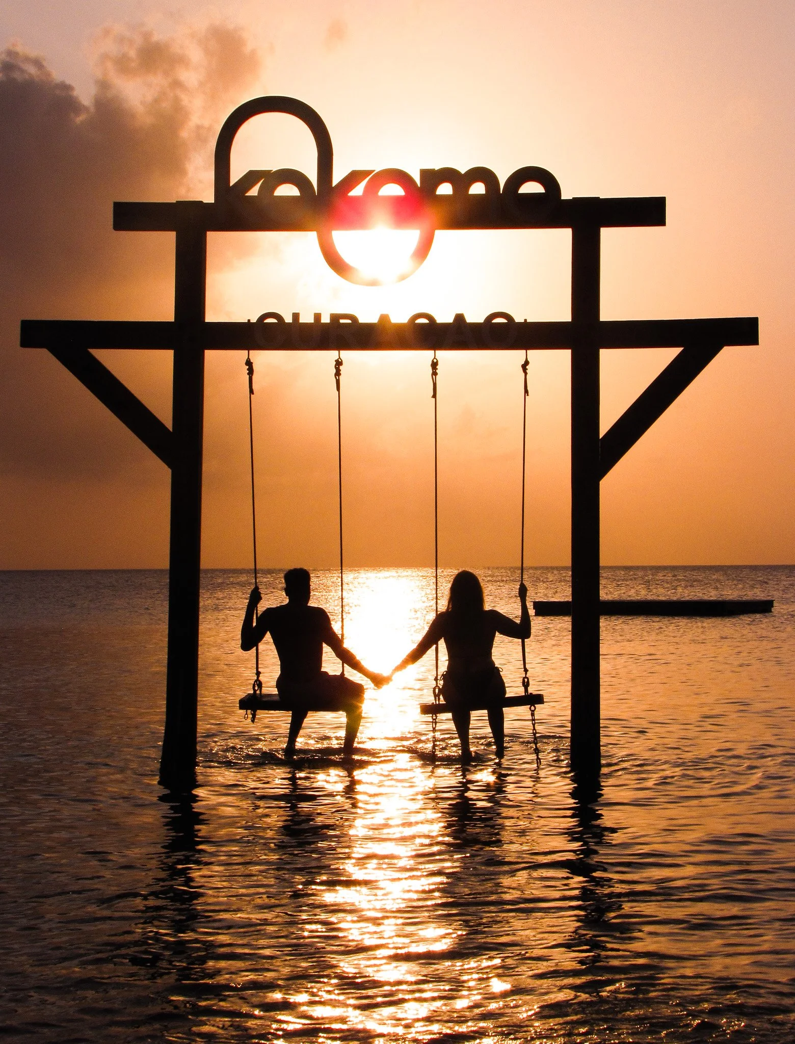Silhouettes of a man and woman sitting on a swing set by the water at sunset, holding hands, with the sun setting over the ocean and the sky with clouds in the background.