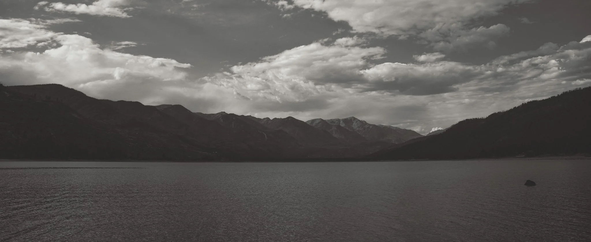 A black and white landscape featuring a body of water in the foreground, mountains in the background, and a sky filled with clouds.