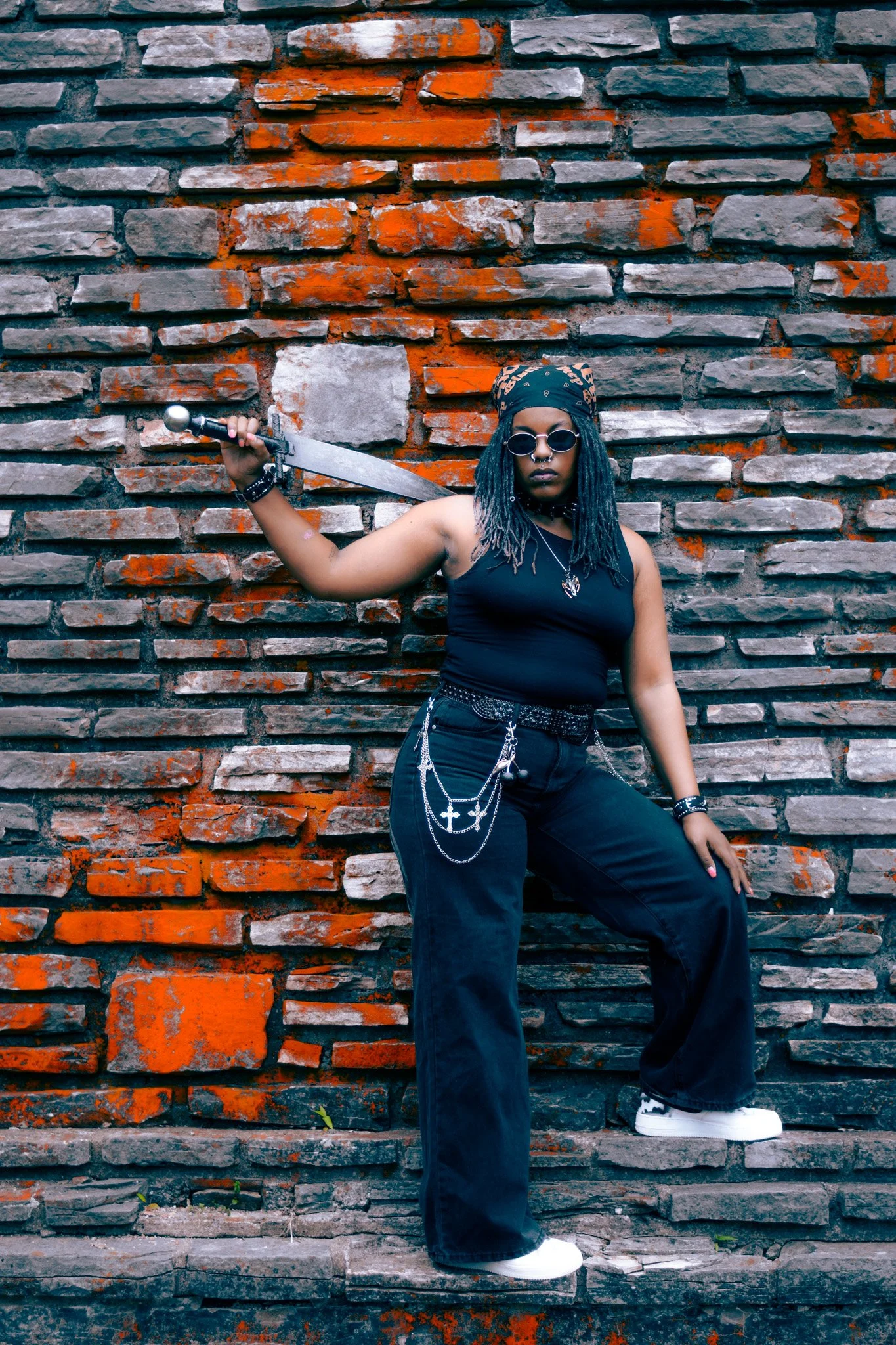 A person with dreadlocks wearing sunglasses, a bandana, dark clothing, chains, and holding a sword standing against a brick wall.