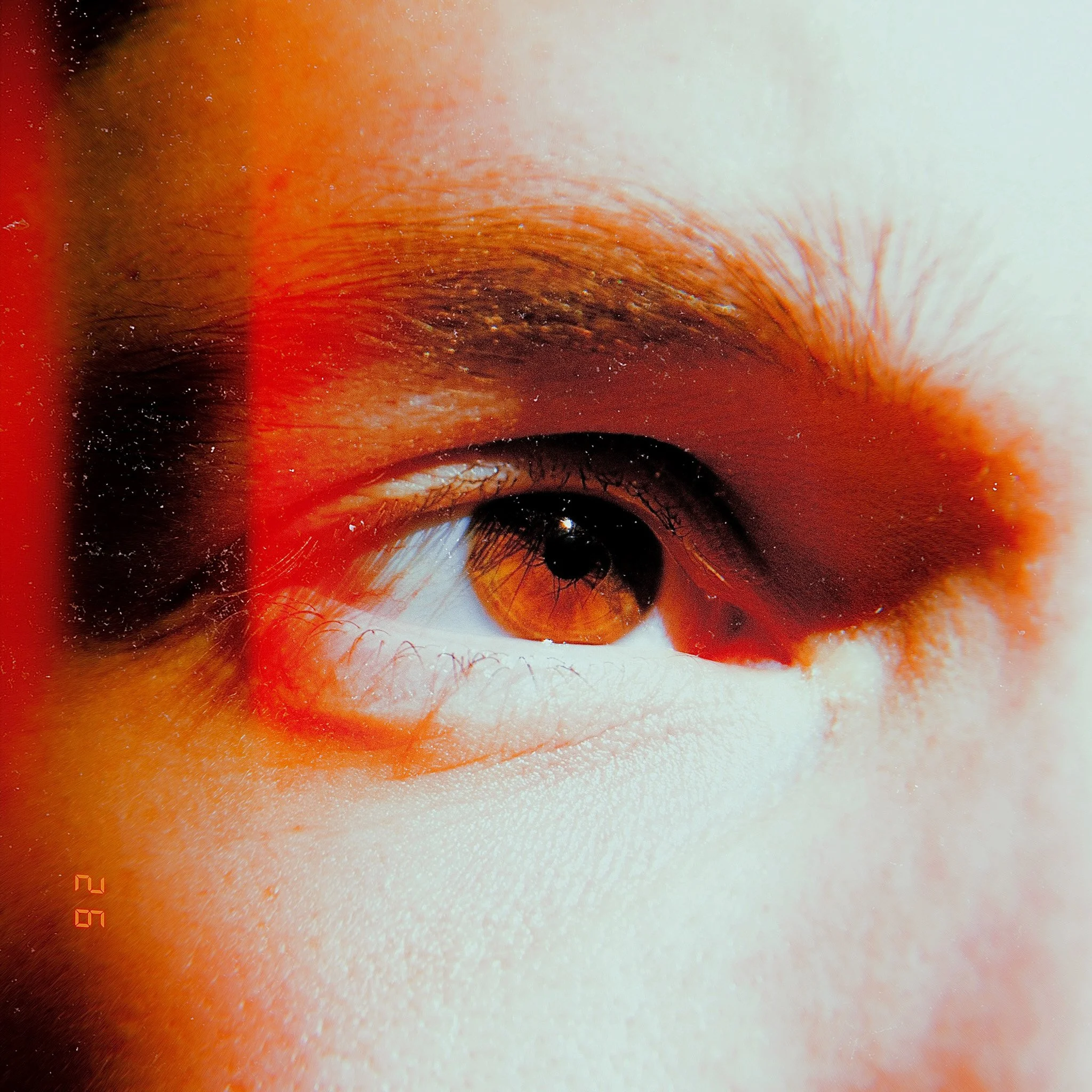 Close-up photograph of a human eye with brown iris, detailed eyelashes, and surrounding skin. The image is magnified and artistic, emphasizing the eye's texture and color.